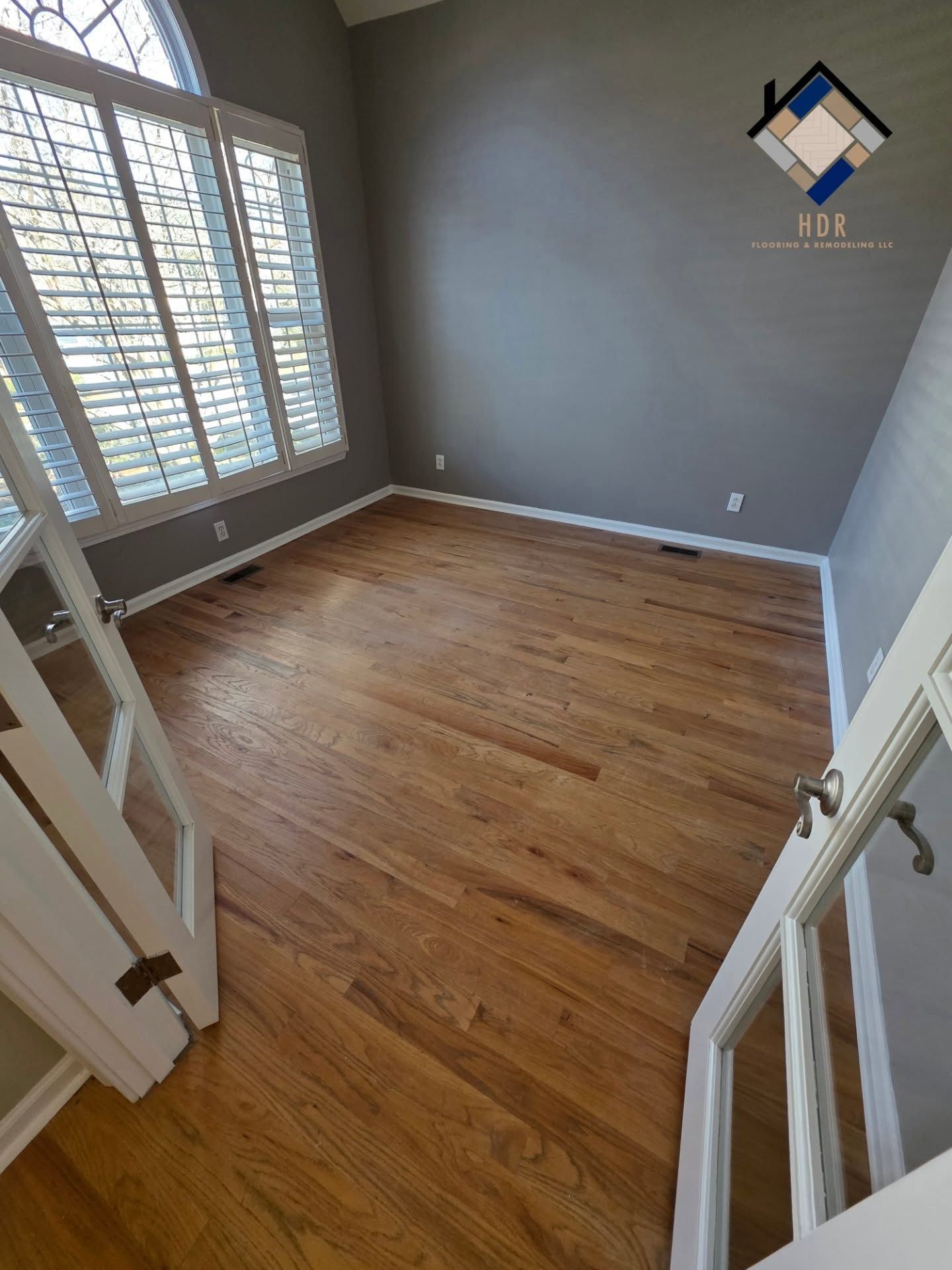 Empty room with hardwood floor, gray walls, and large window shutters, viewed from the doorway