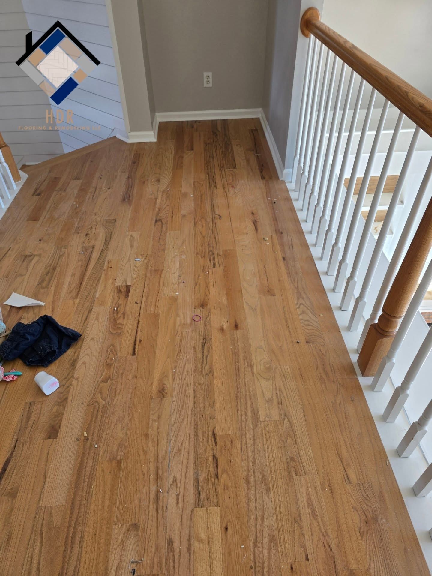 Narrow hardwood hallway with white railing, tan walls, and a few items on the floor
