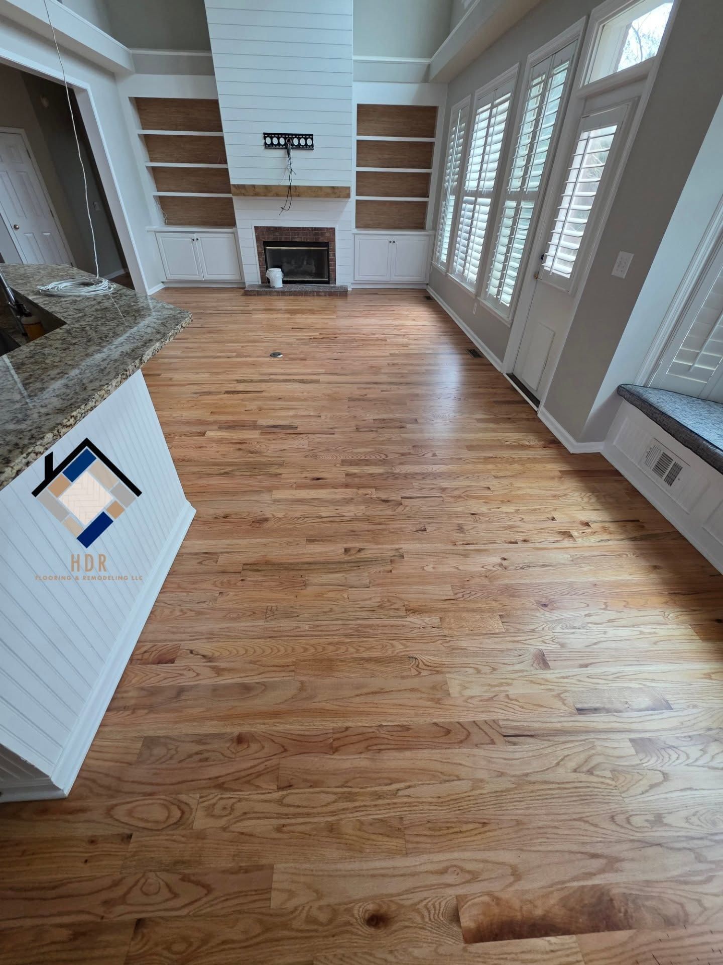 Empty living room with hardwood floors, fireplace, built-in shelves, and large windows with shutters.