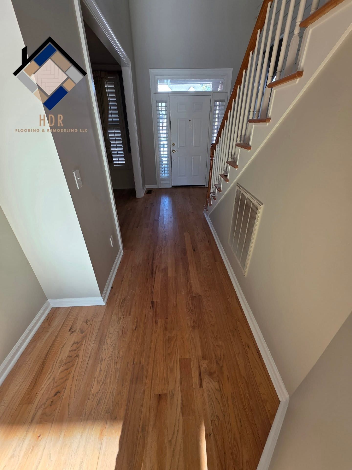 Hallway with wood floors, staircase railing, front door, and sunlight streaming in from the left.