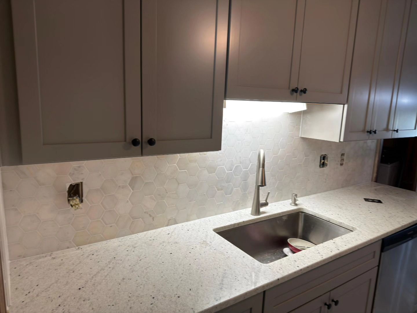 Kitchen countertop with gray cabinets, white backsplash, and stainless steel sink under cabinet lighting