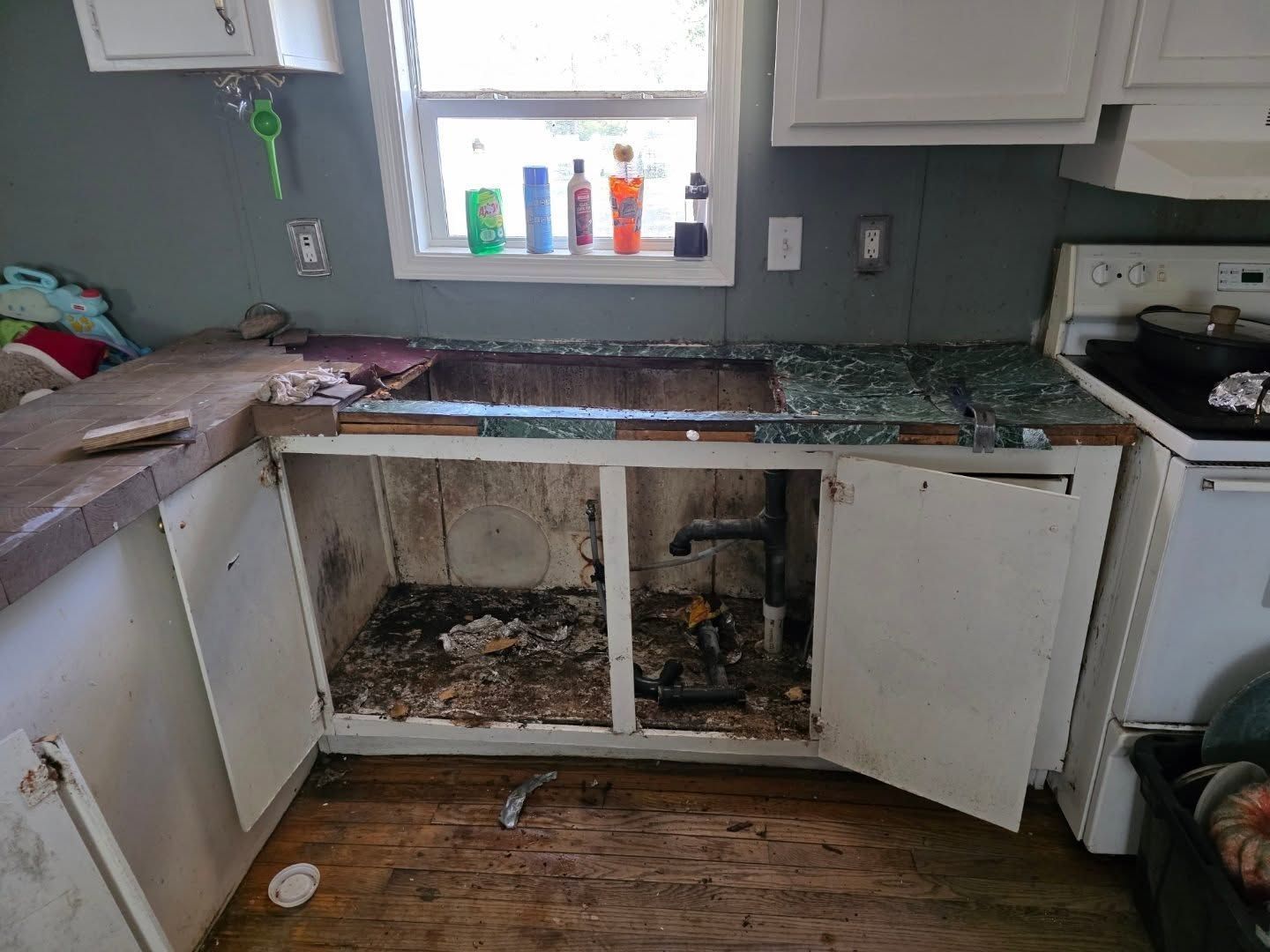 Damaged kitchen with broken cabinets, exposed under-sink area, and debris on the floor.