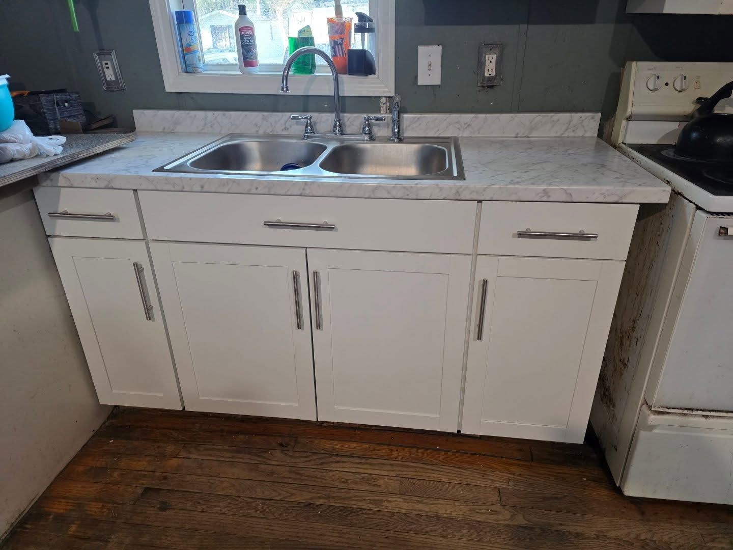 White kitchen sink cabinet with double basin, countertop, and wood floor beside a stove and refrigerator.
