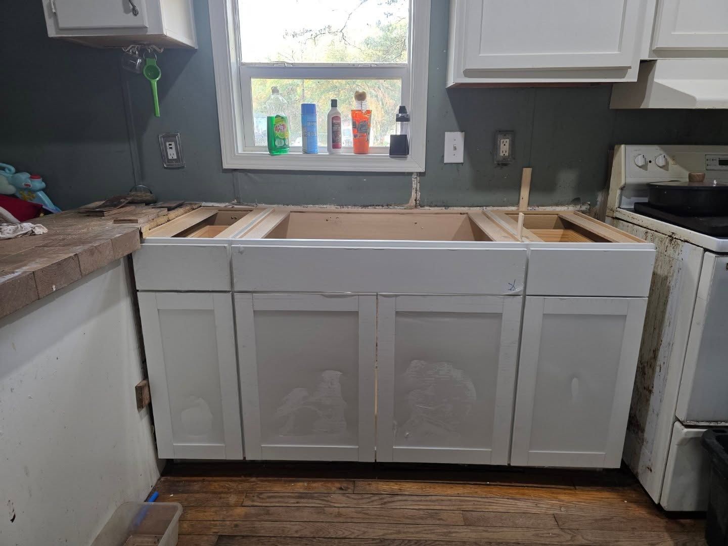 Kitchen under renovation with white cabinets, exposed countertop framing, and a window above the sink.