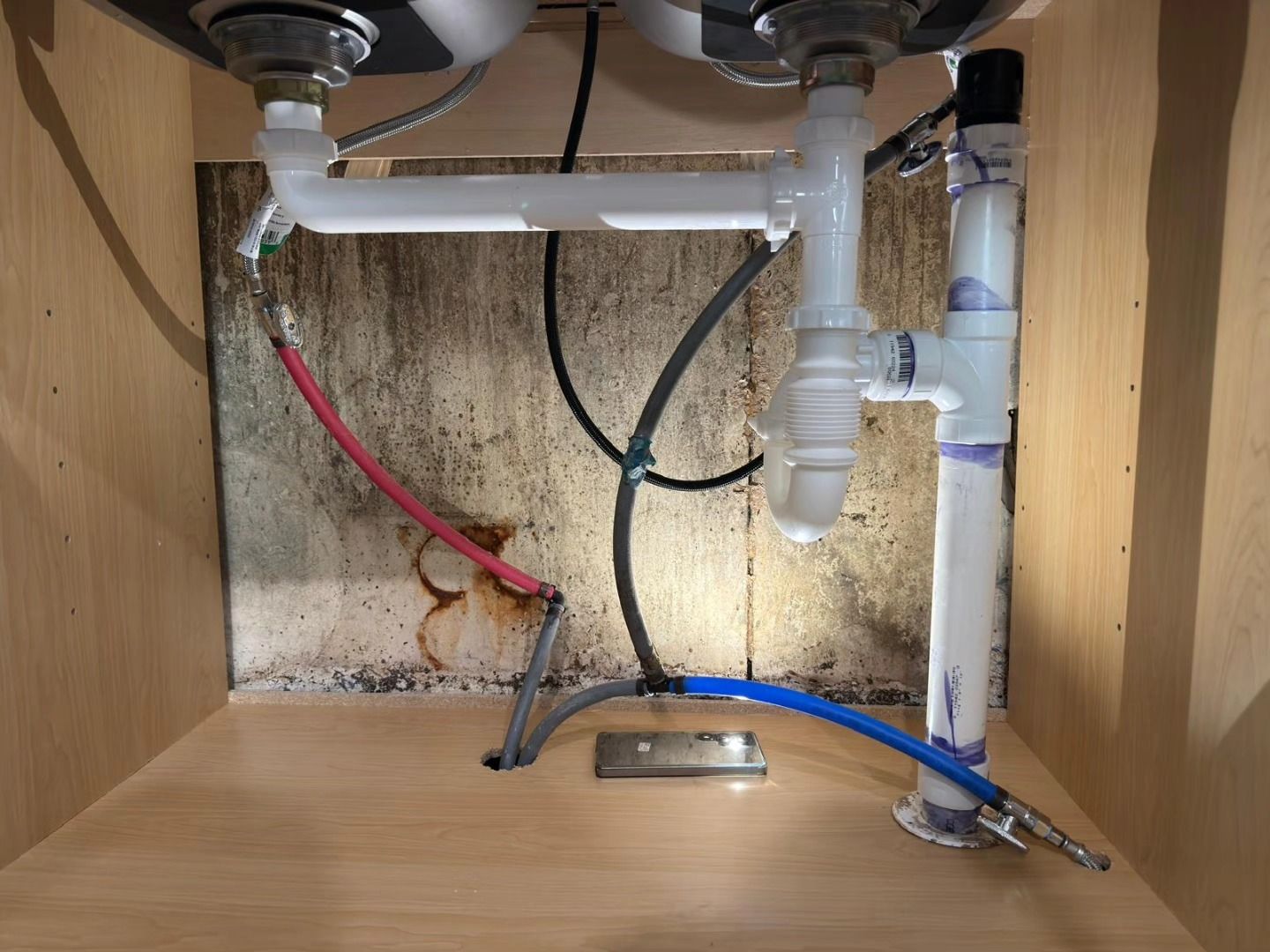 Under-sink plumbing with red and black wires, white drain pipes, and a blue hose in a wooden cabinet.