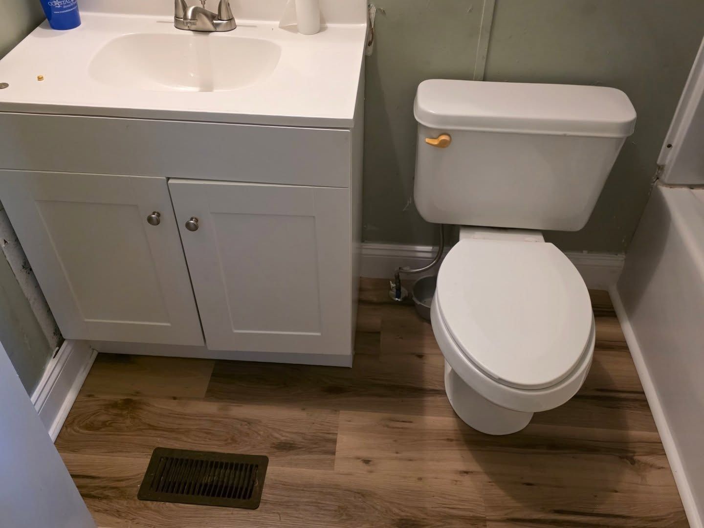 Small bathroom with white vanity and toilet, wood-look floor, and wall vent.