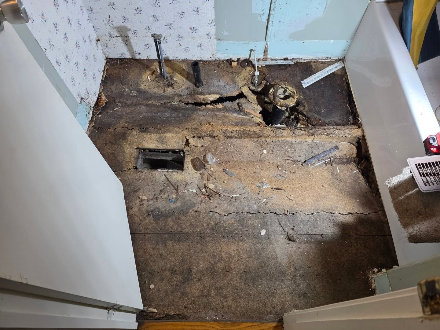 Exposed bathroom subfloor with plumbing holes and debris during renovation