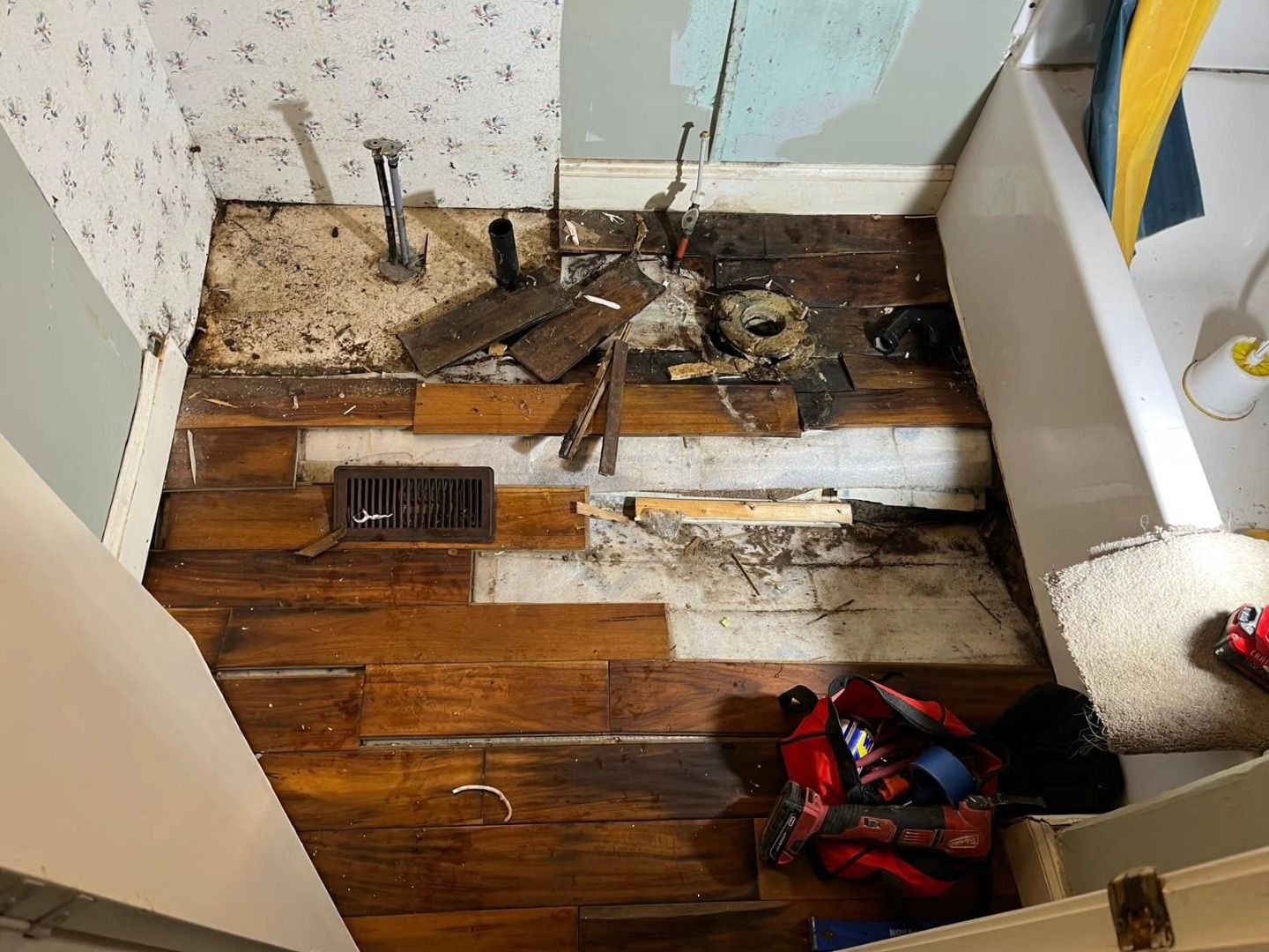 Flood-damaged room with torn-up floorboards, exposed subfloor, debris, and a red bag in the corner