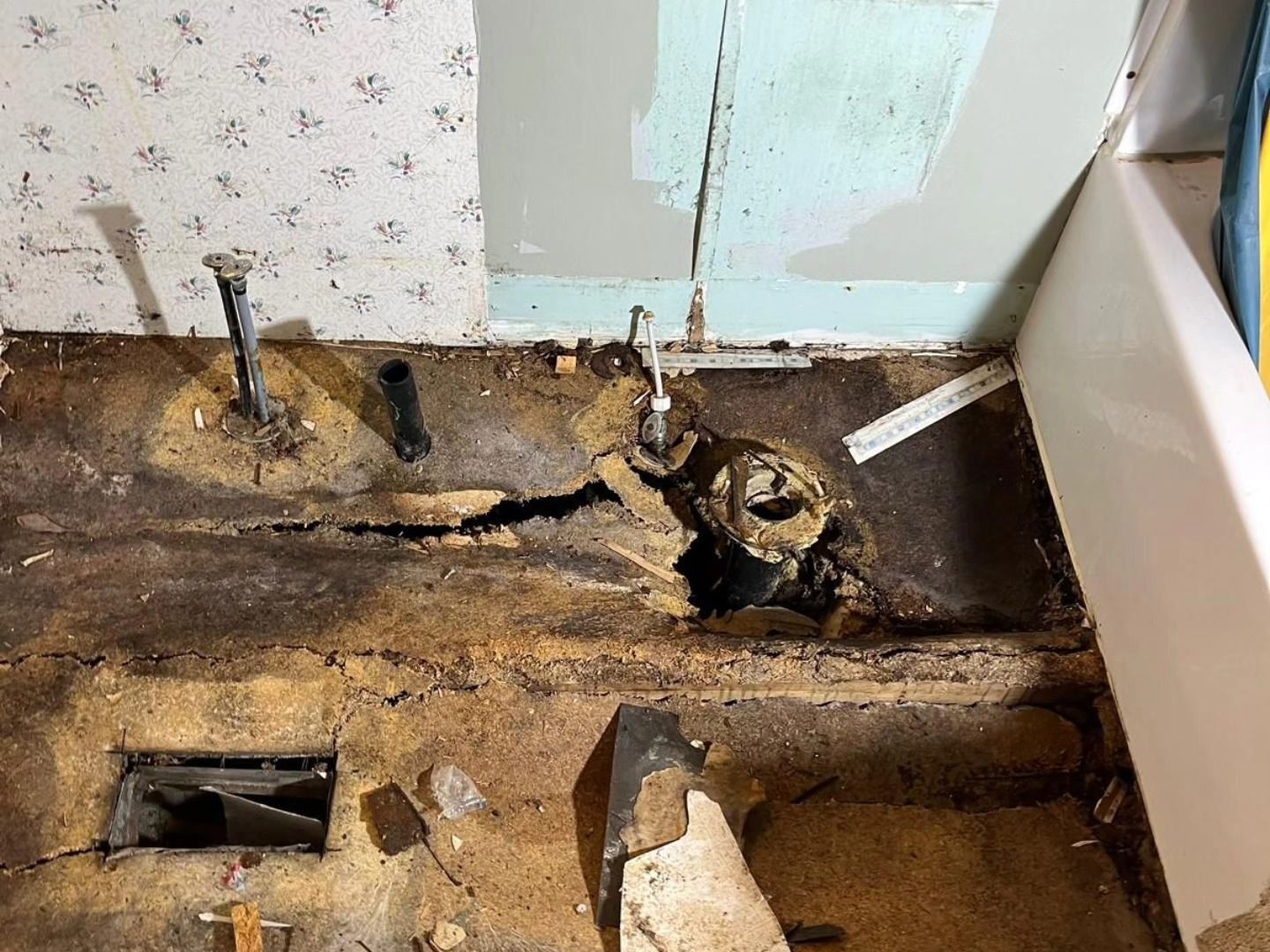 Damaged bathroom floor with exposed pipes and a broken, rotten section near a wall and tub