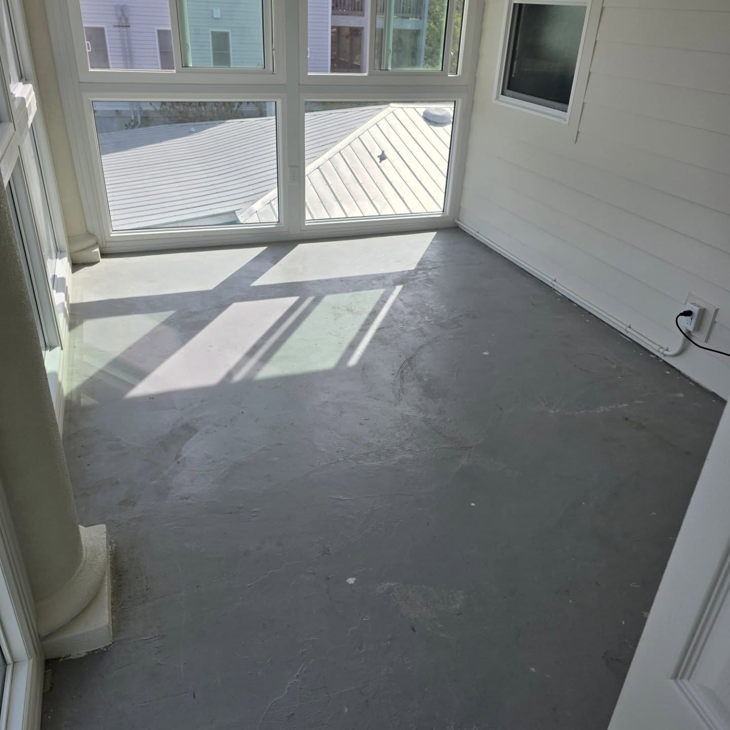 Empty sunroom with gray concrete floor and large windows casting sunlight and shadows.