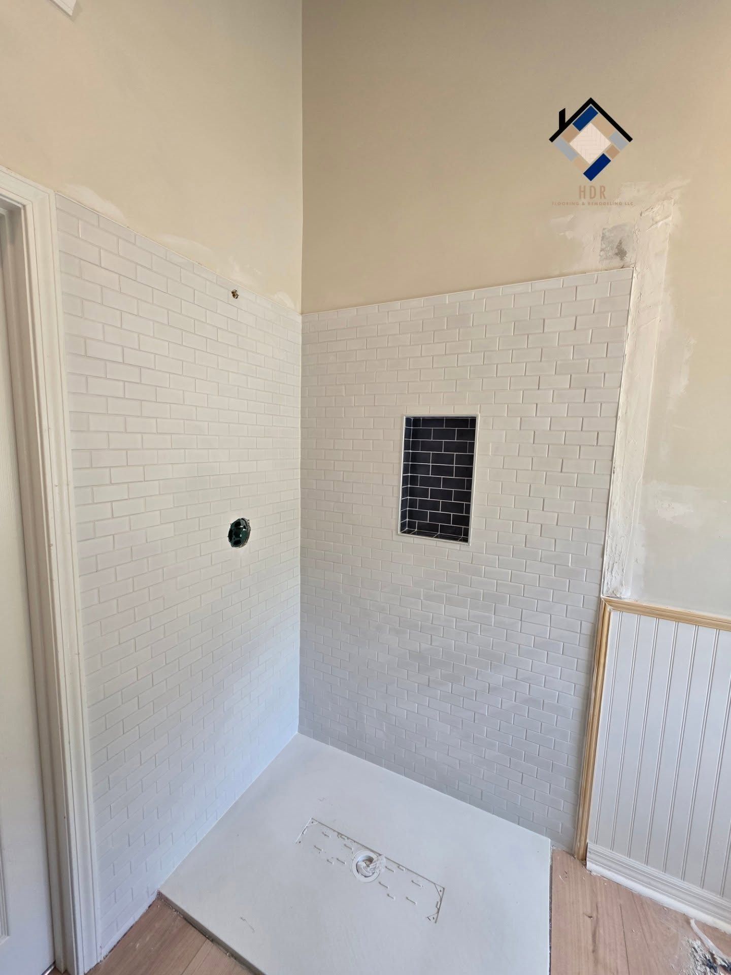 Tiled shower under renovation with white wall tiles, a small dark tile panel, and a white shower base