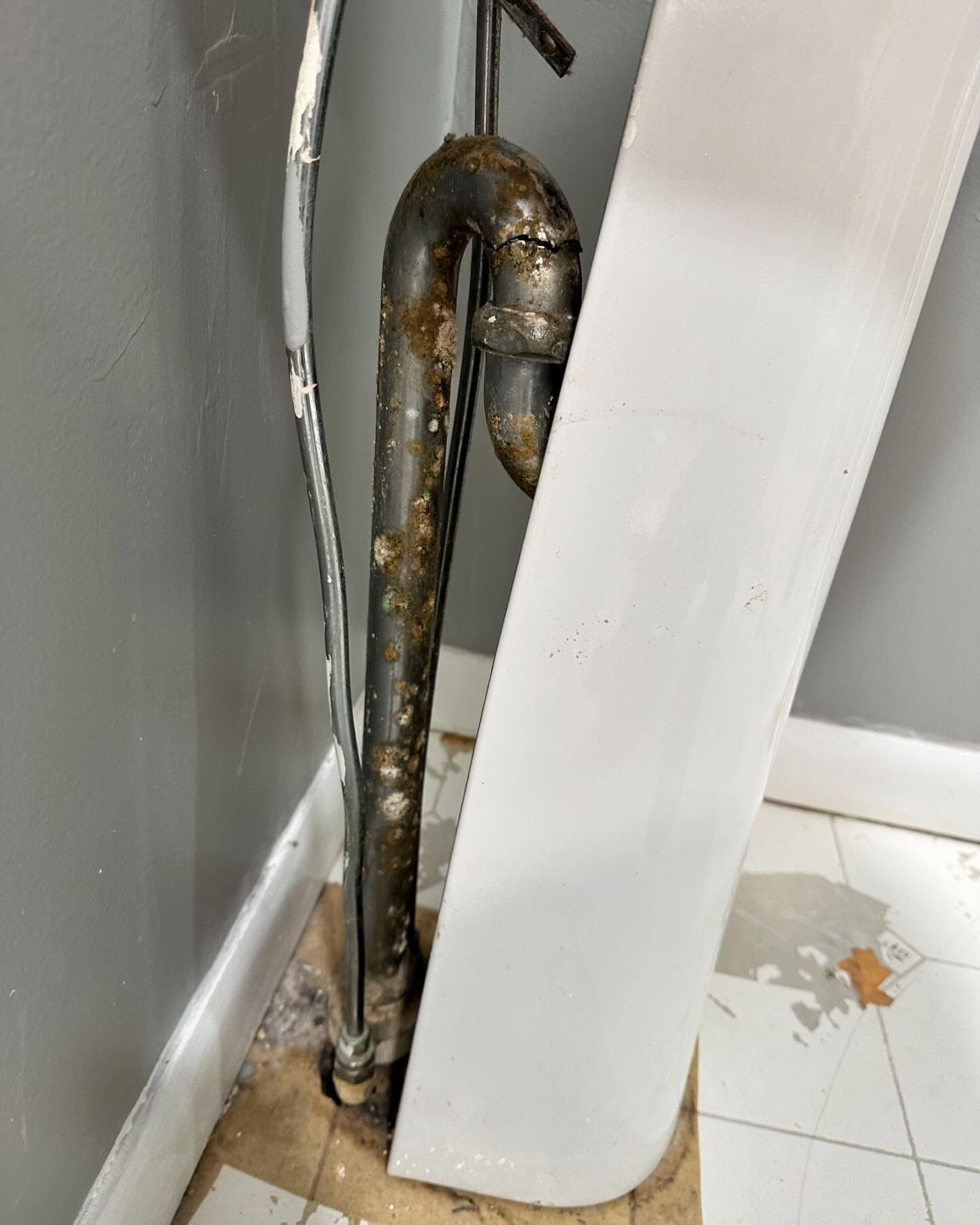 Rusty metal pipe behind white panel on a gray wall, with water stains on the floor.