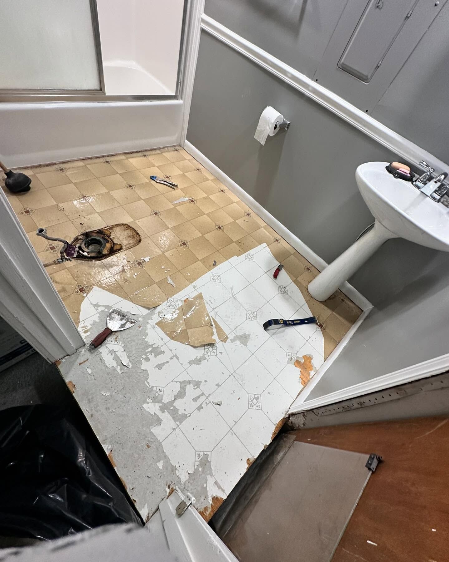 Damaged bathroom floor with torn white tiles, scattered debris, and a sink beside a shower area