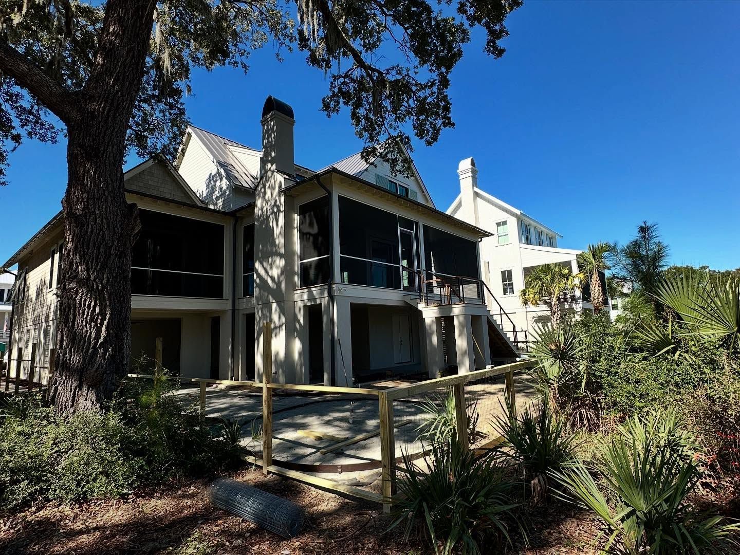 Two-story house with dark trim and balconies, viewed through trees and landscaping on a sunny day
