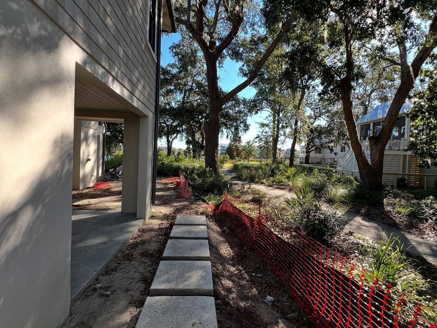 Side yard walkway beside a house with concrete pavers, red netting, trees, and a garden bed in sunlight