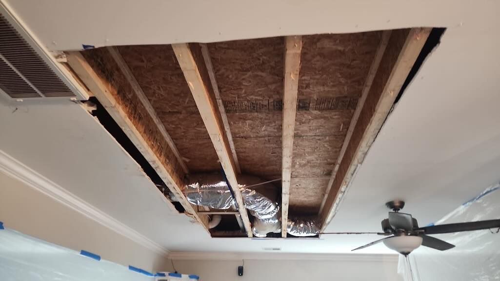 Ceiling with a large rectangular section cut out, exposing wooden joists and ductwork in a room with a fan below
