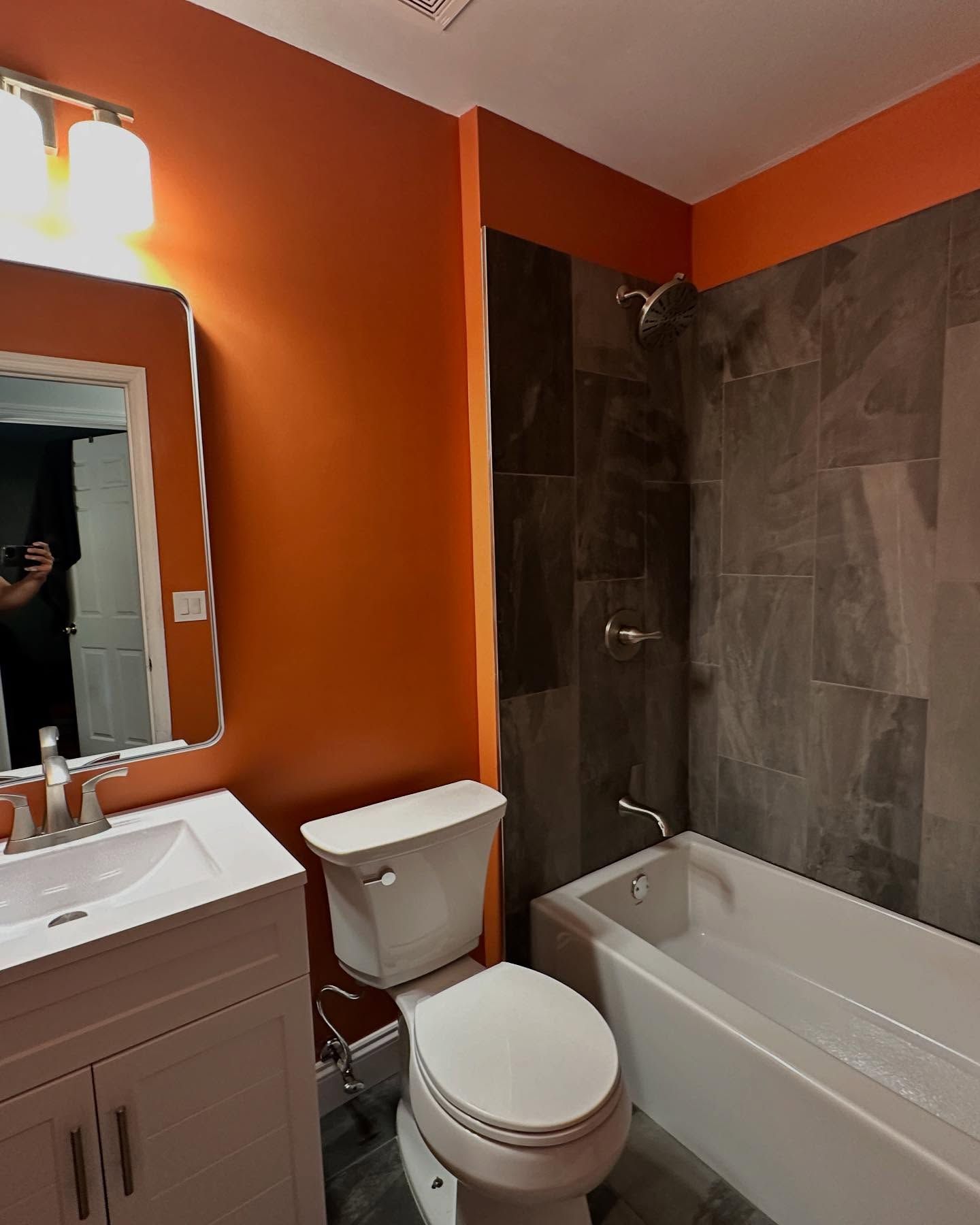 Small bathroom with orange walls, white sink and toilet, and a bathtub with dark tile surround.