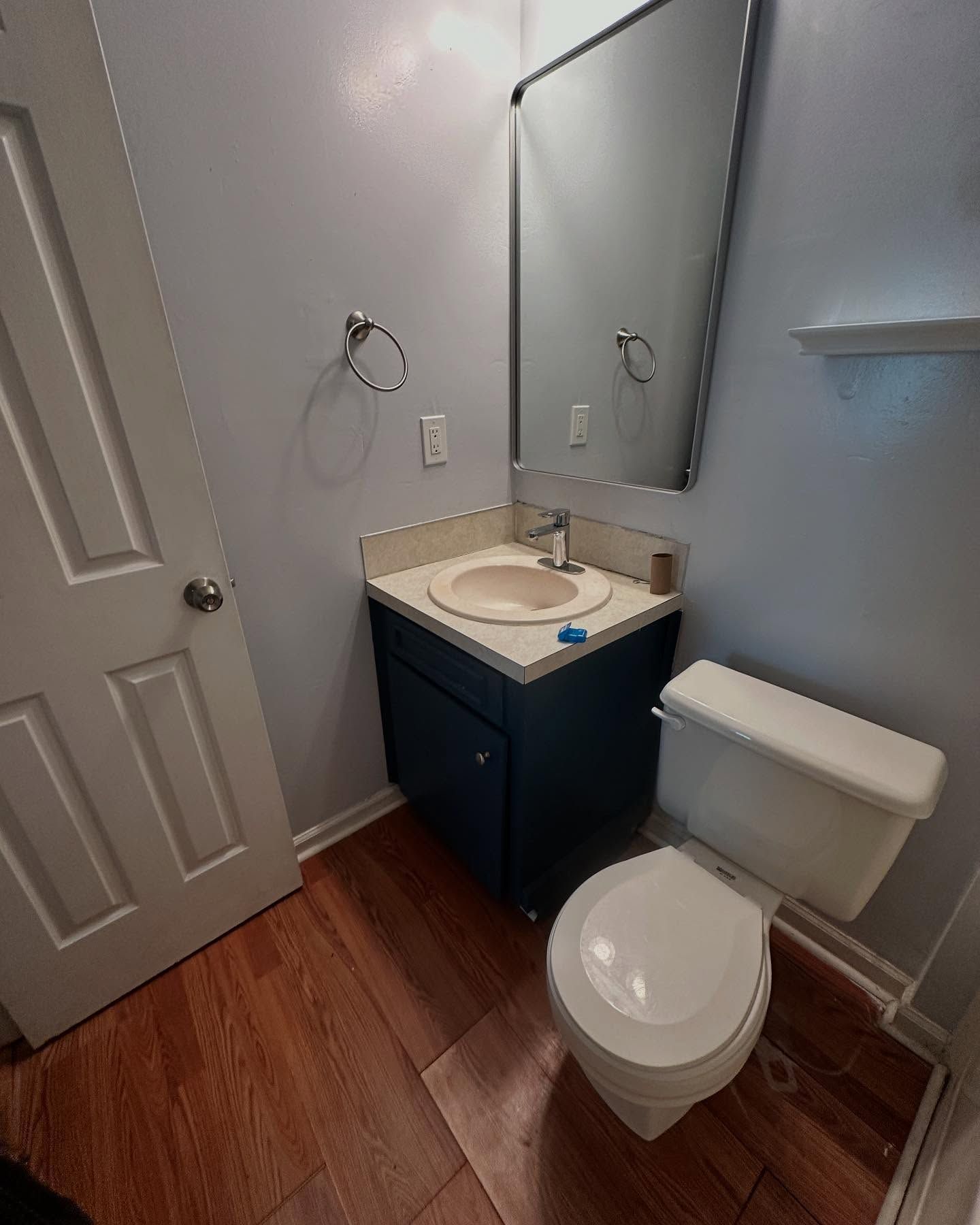 Small bathroom with toilet, sink vanity, mirror, and white door on wood-look floor.