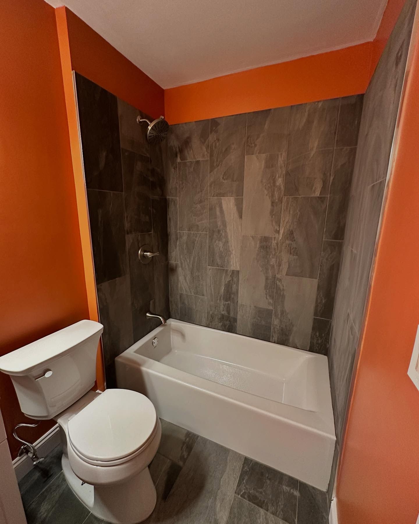 Small bathroom with orange walls, white toilet and tub, and gray tile shower surround.
