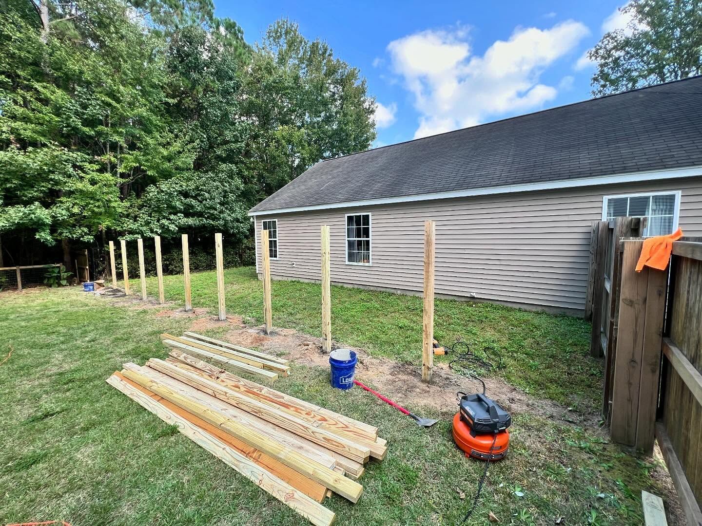 Backyard fence under construction beside a house, with posts, lumber, a bucket, and a saw on the grass.