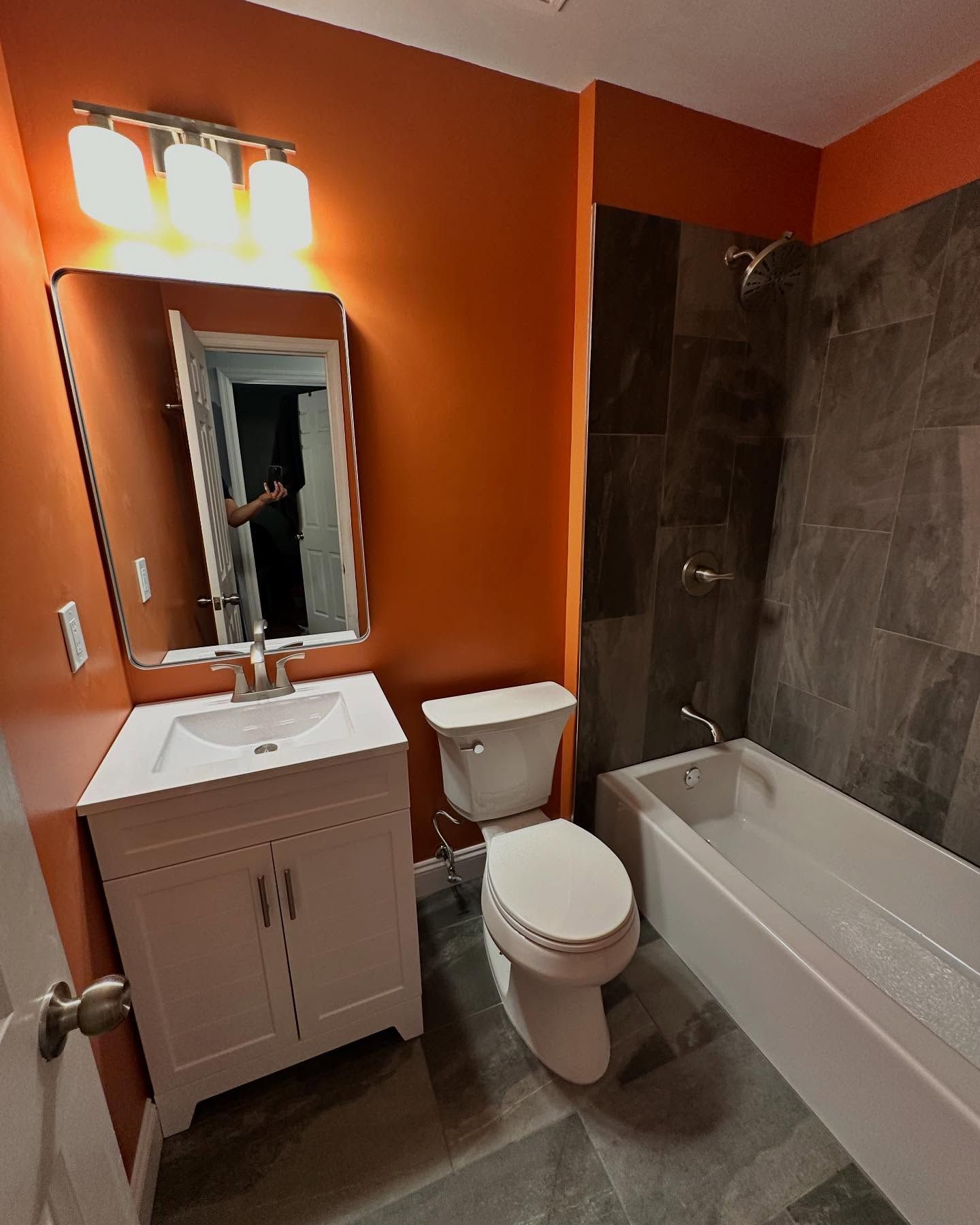 Small bathroom with orange walls, white vanity and toilet, and a tiled bathtub/shower.