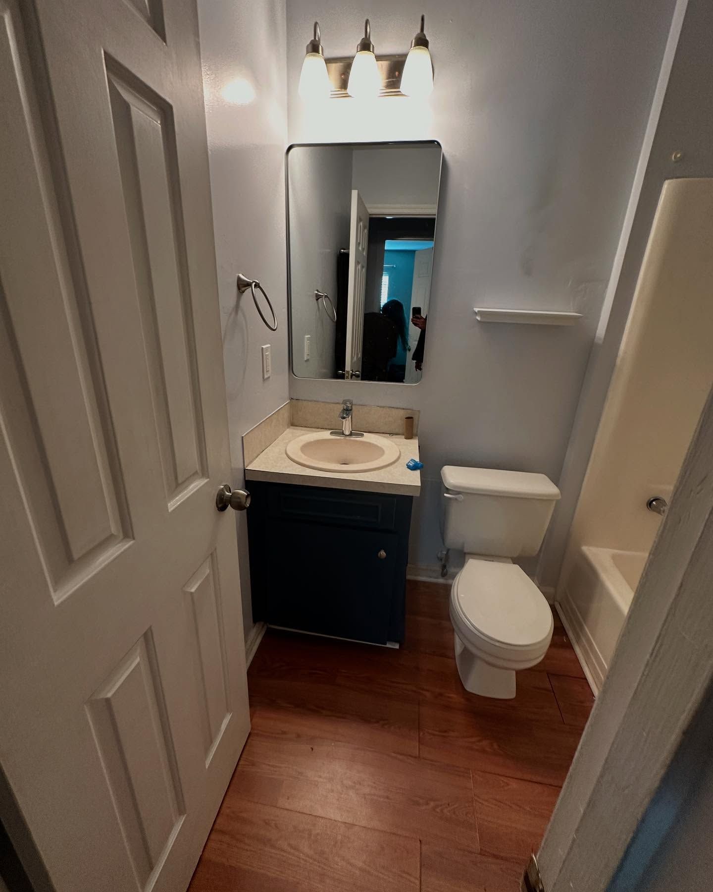 Small bathroom with a sink, mirror, toilet, and wood floor in a narrow white-walled space