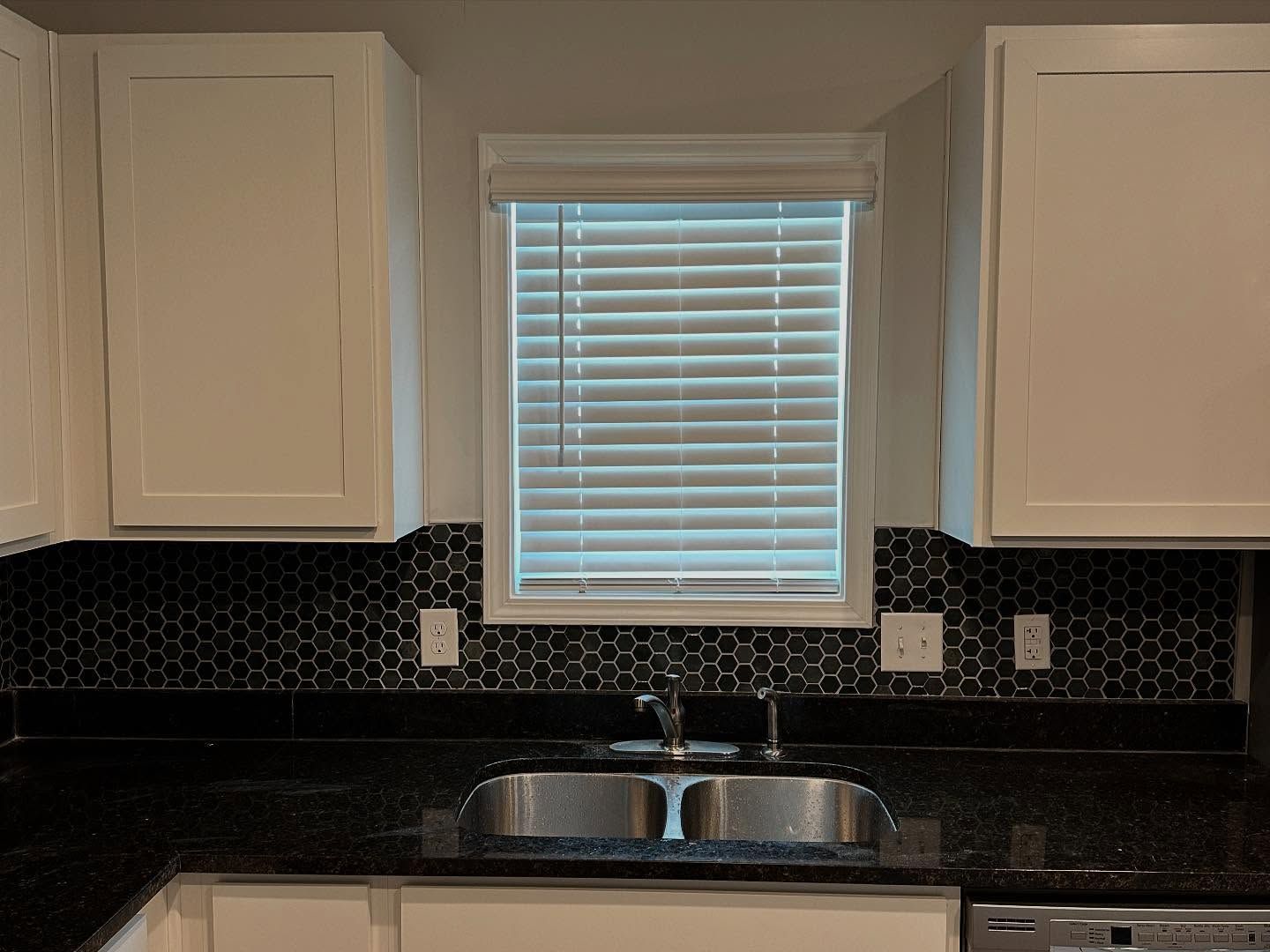 Kitchen sink under a window, with white cabinets, black tile backsplash, and dark countertop.