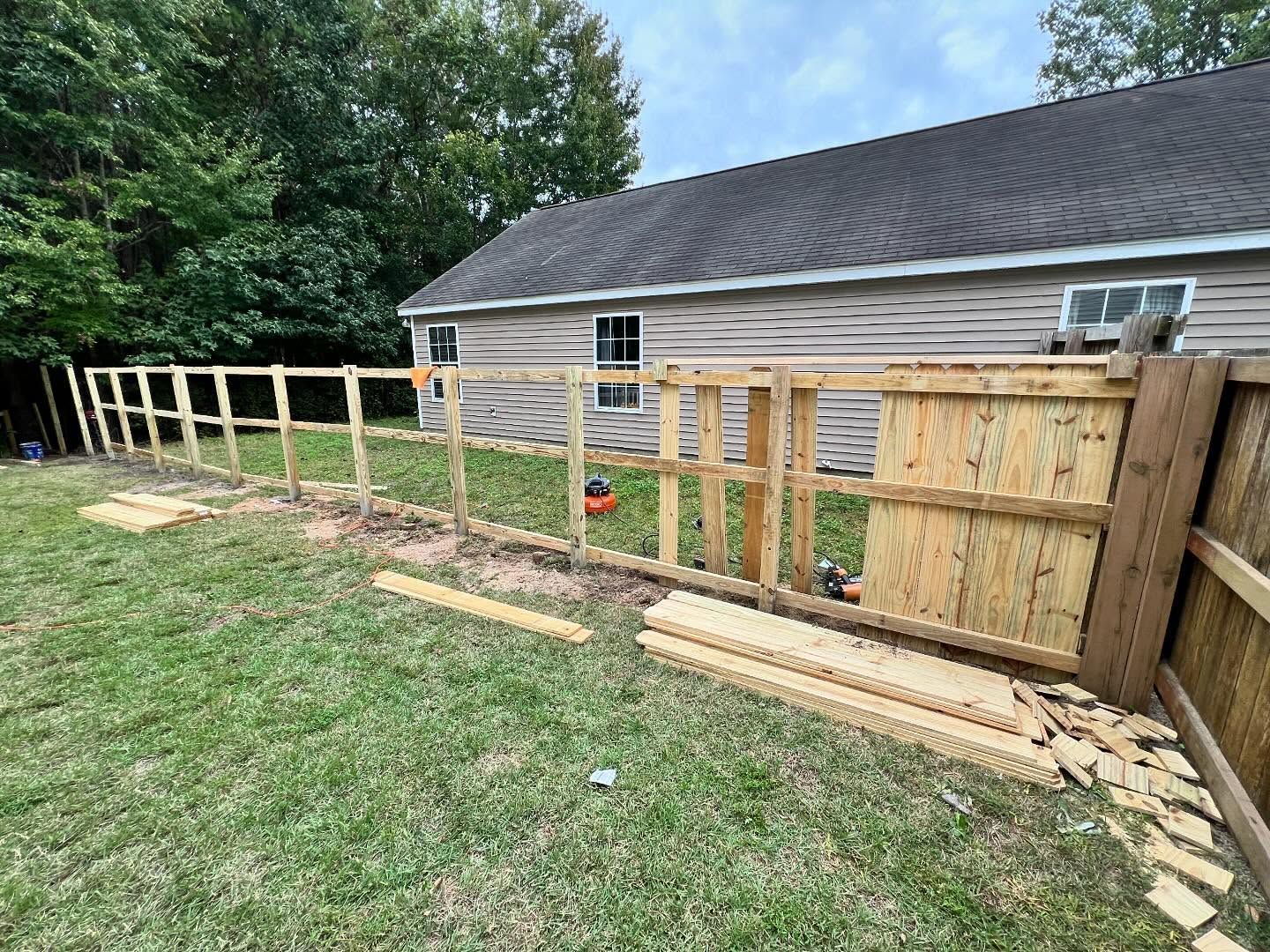Wooden fence under construction along a house beside a grassy yard, with posts, panels, and lumber laid out.