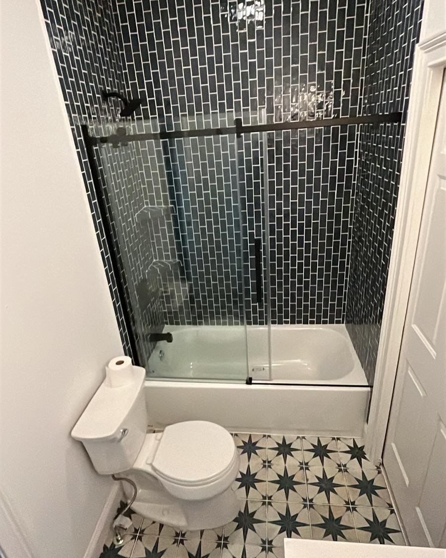 Small bathroom with toilet, glass shower, black mosaic tile wall, and patterned floor tiles.