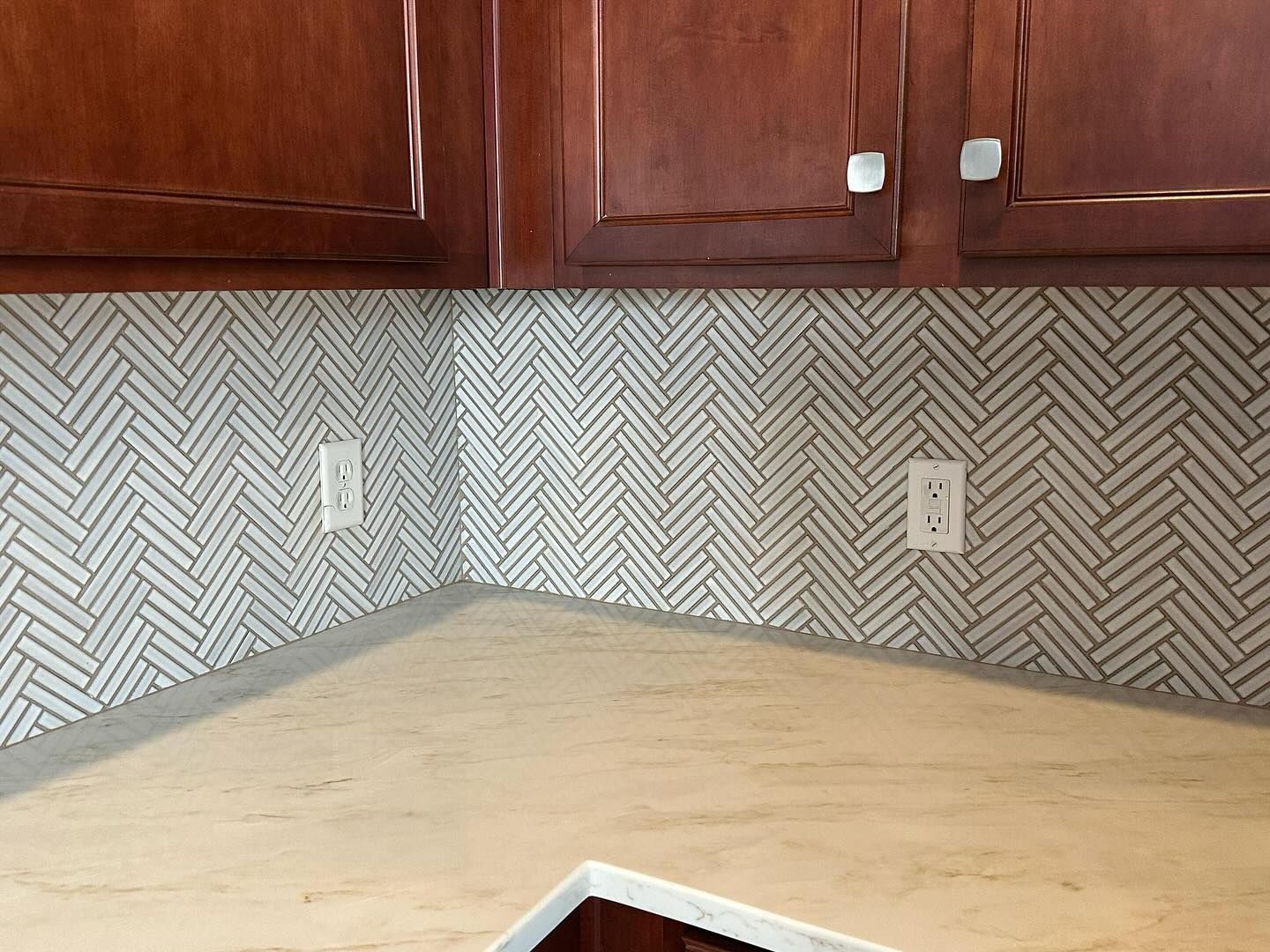 Kitchen corner with wood cabinets, gray chevron backsplash, beige countertop, and electrical outlets