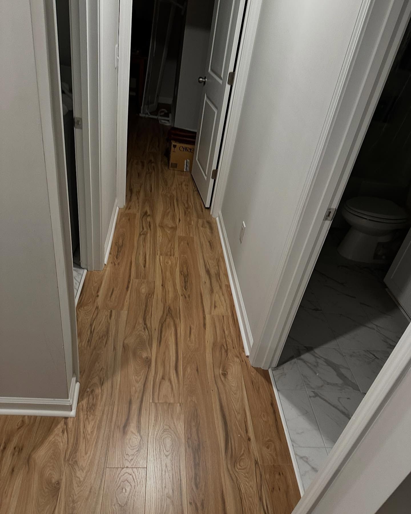 Narrow hallway with wood flooring, white walls, and open doorways leading to a bathroom and dim room.
