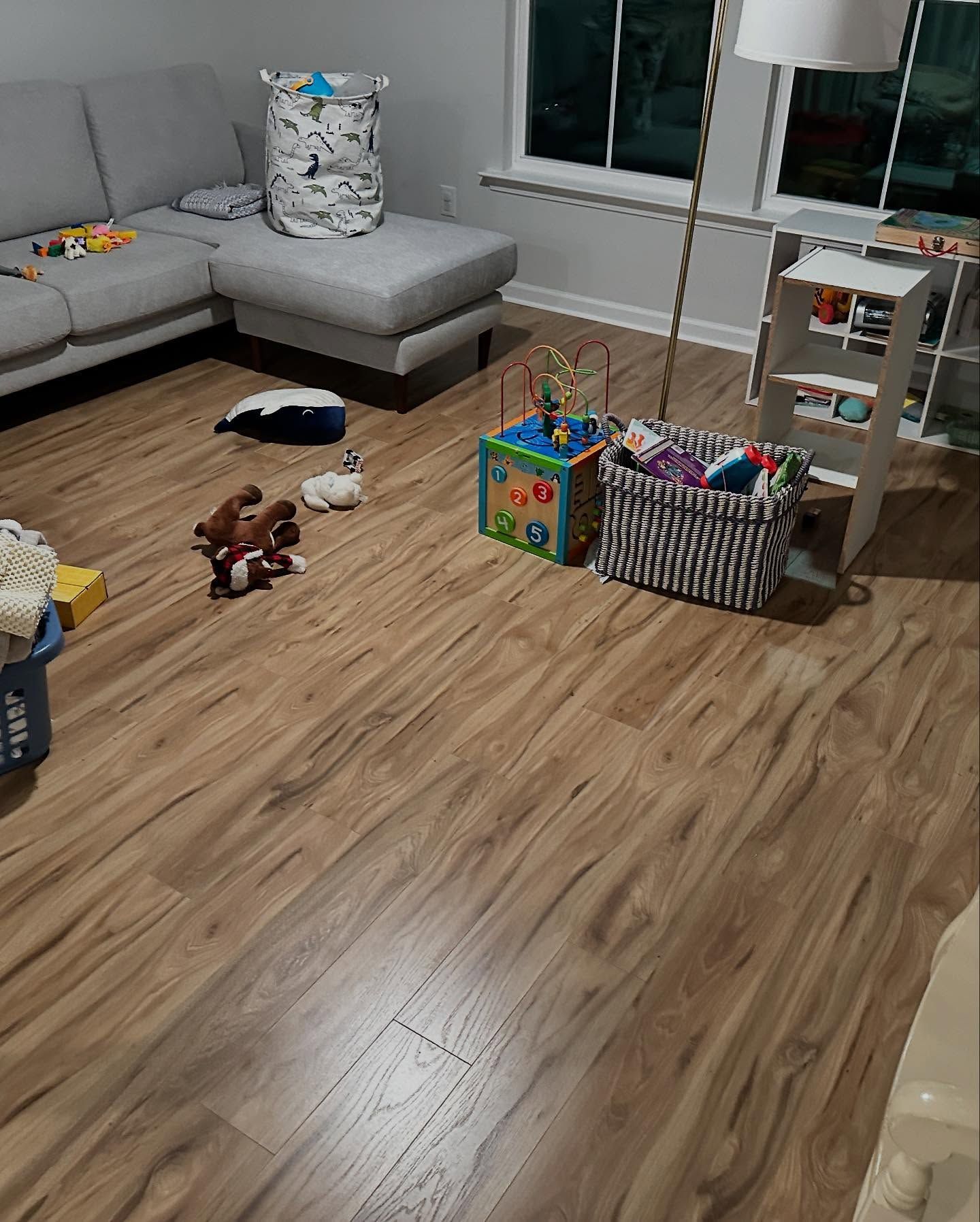 Living room with gray sectional, toys scattered on wood floor, and a dining table near the window.