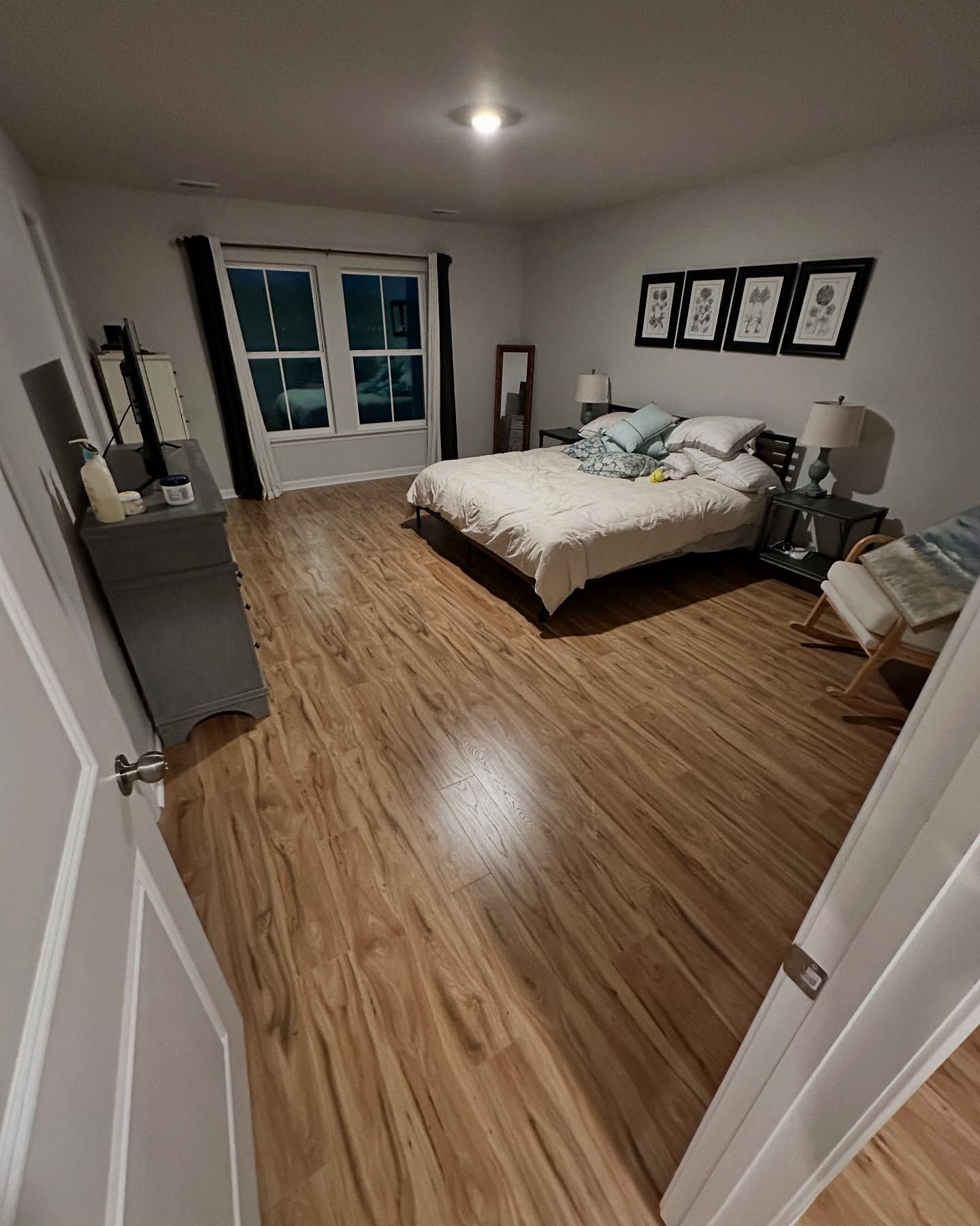Spacious bedroom with wood floors, white bed, dark dresser, and framed wall art near a window.