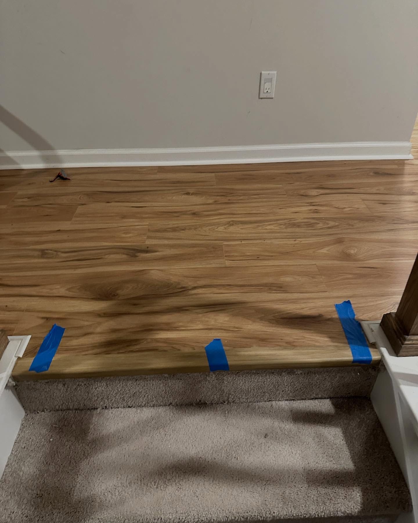 Indoor wood floor at top of carpeted stairs, with blue tape marking the edge and a wall outlet ahead.