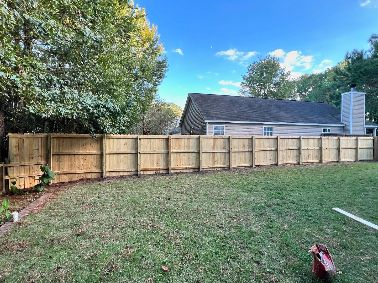 Backyard with grass, wooden fence, trees, and a house under a blue sky