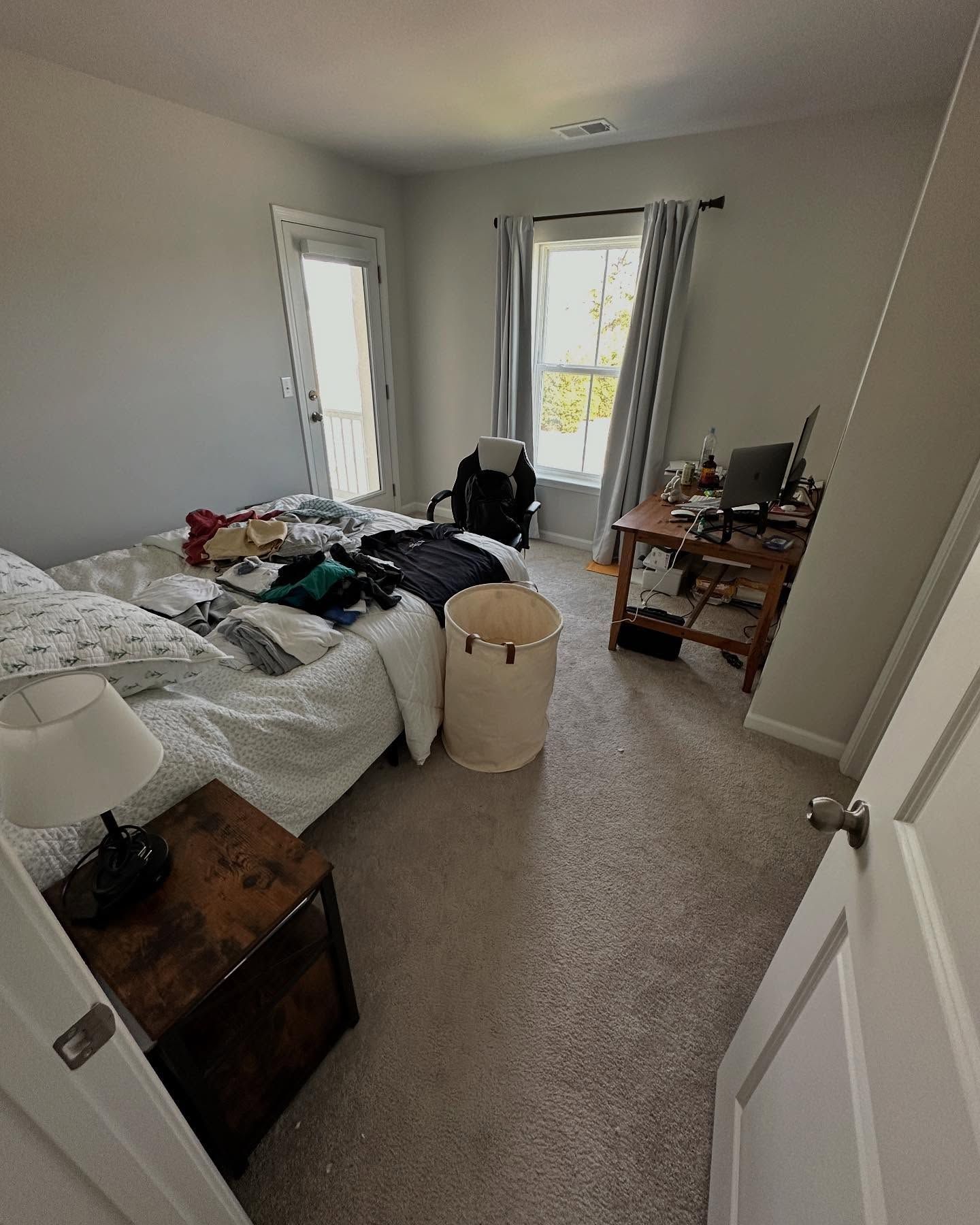 Messy bedroom with unmade bed, suitcase, desk, and open balcony doors letting in daylight