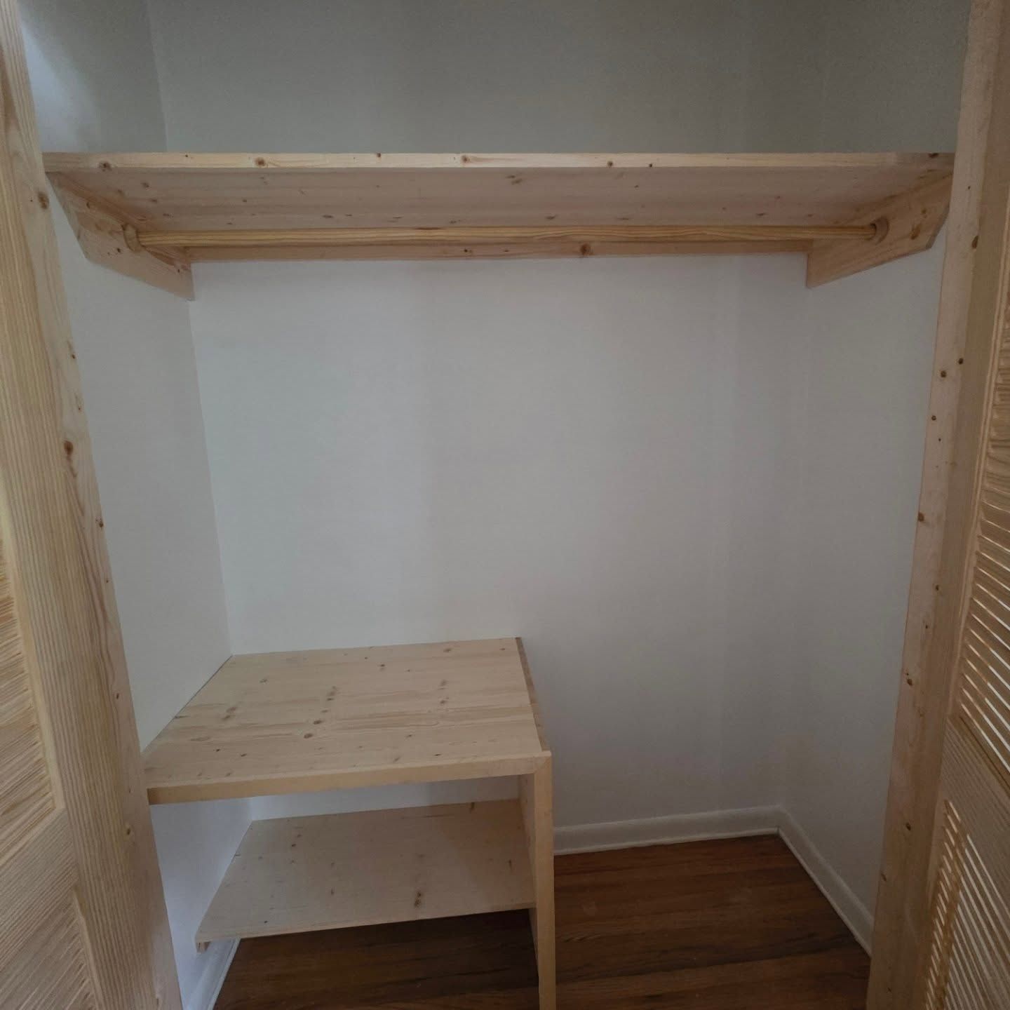 Small walk-in closet with white walls, wood floor, built-in shelf, and a low wooden bench.