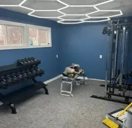 Small gym room with dumbbells, cable machine, bench, and blue walls under hexagonal ceiling lights.