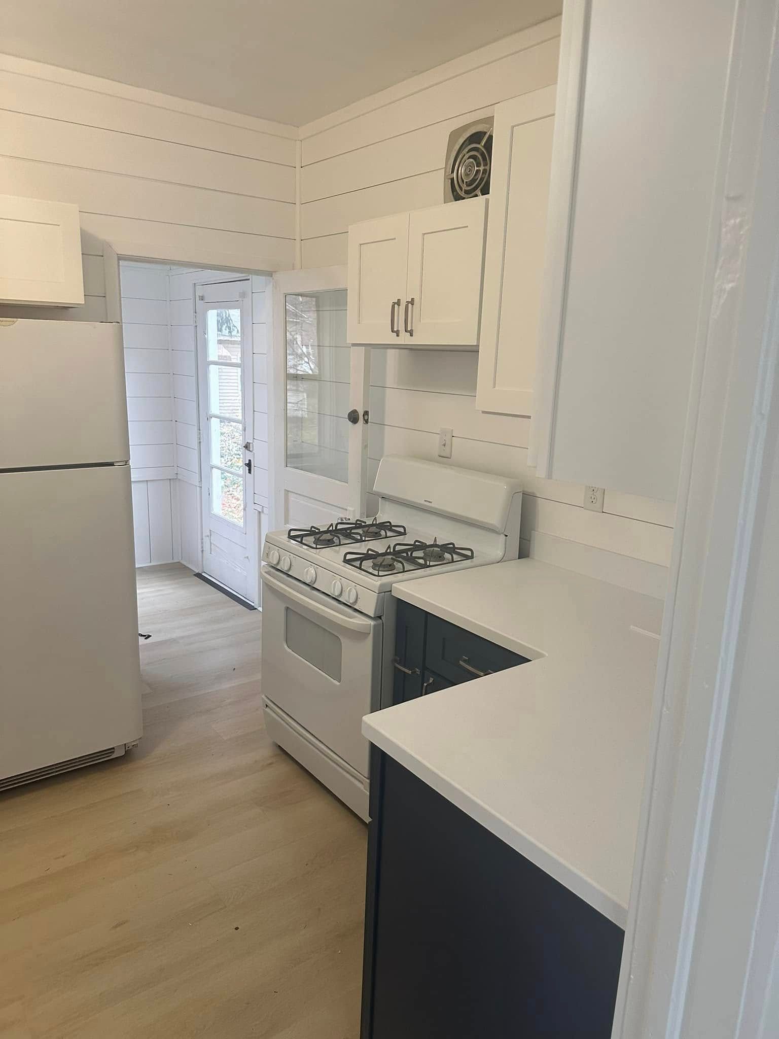 Bright white galley kitchen with stove, refrigerator, cabinets, and light wood flooring.