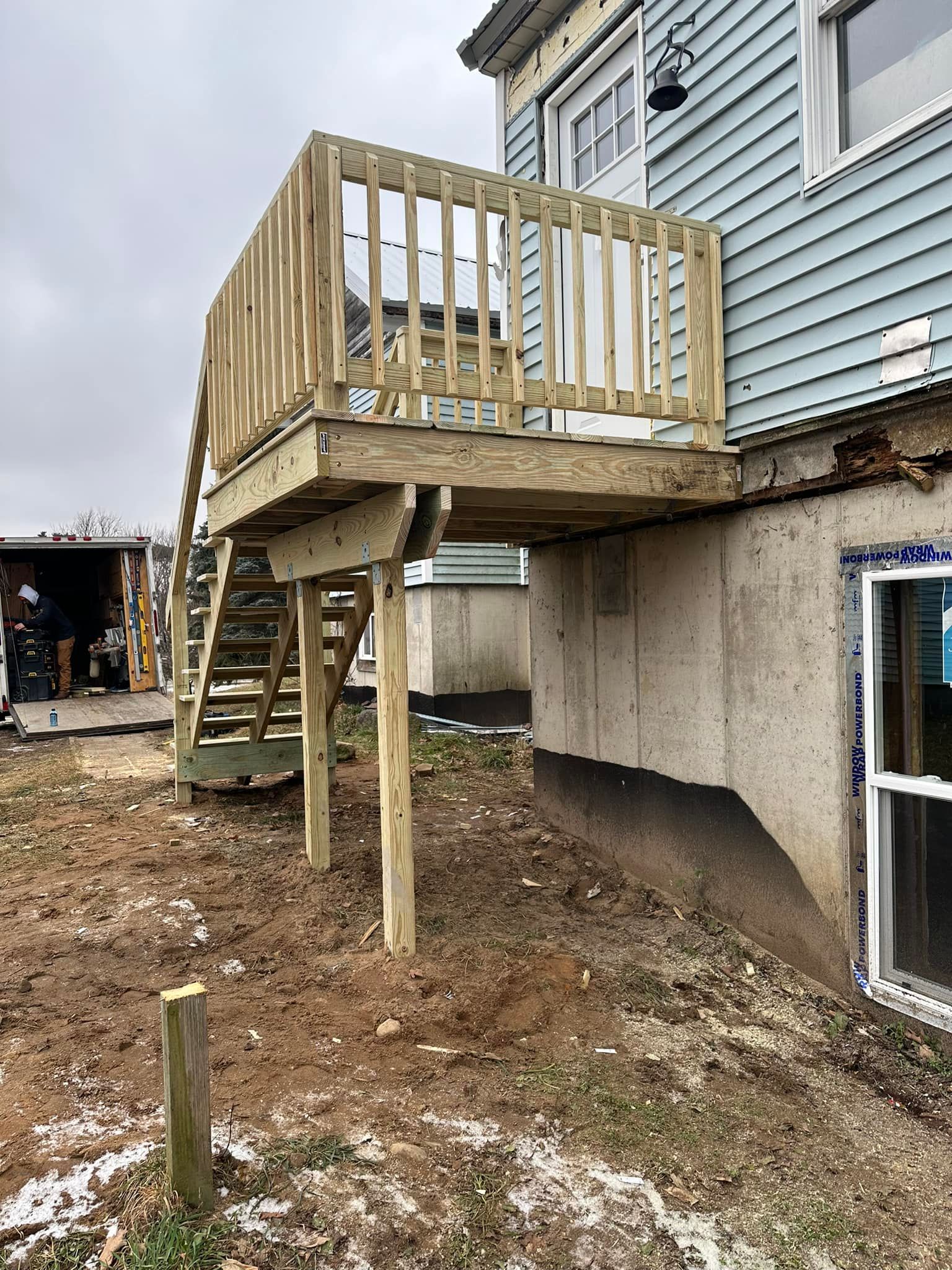 Raised wooden deck under construction beside a house with unfinished support posts and dirt ground.