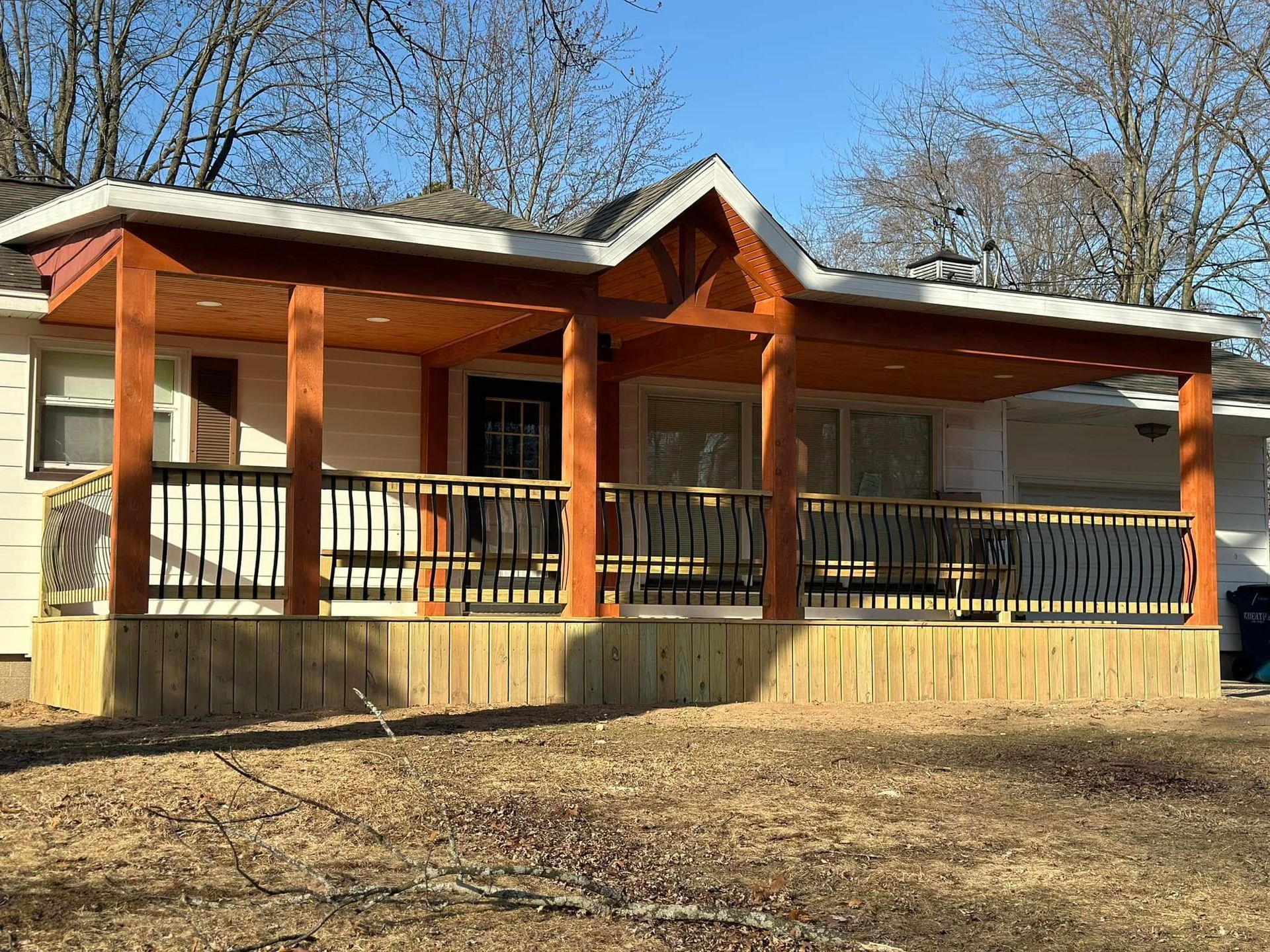 Small wooden house with a covered porch, black railings, and bare trees in the yard.