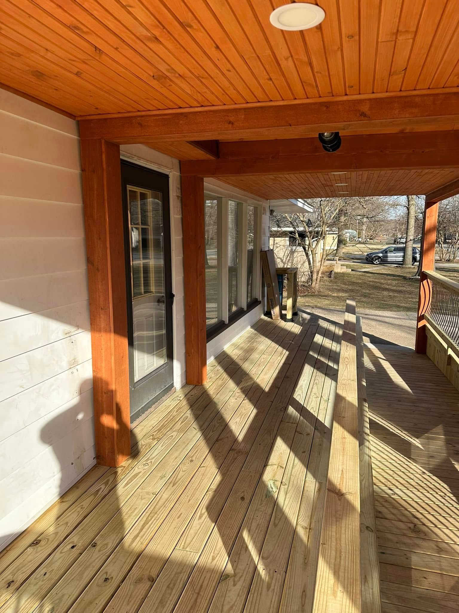 Covered wooden porch with sunlit railing shadows, windows, and a view of a yard beyond