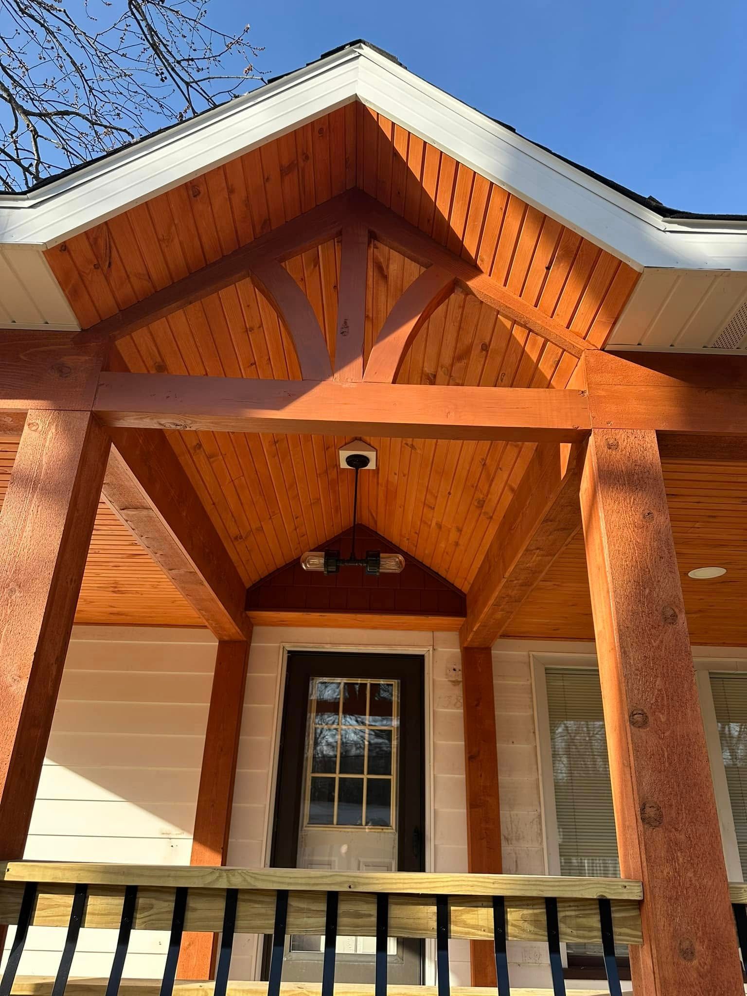 Front porch with wooden gable roof, white trim, tan siding, and black railing under a blue sky