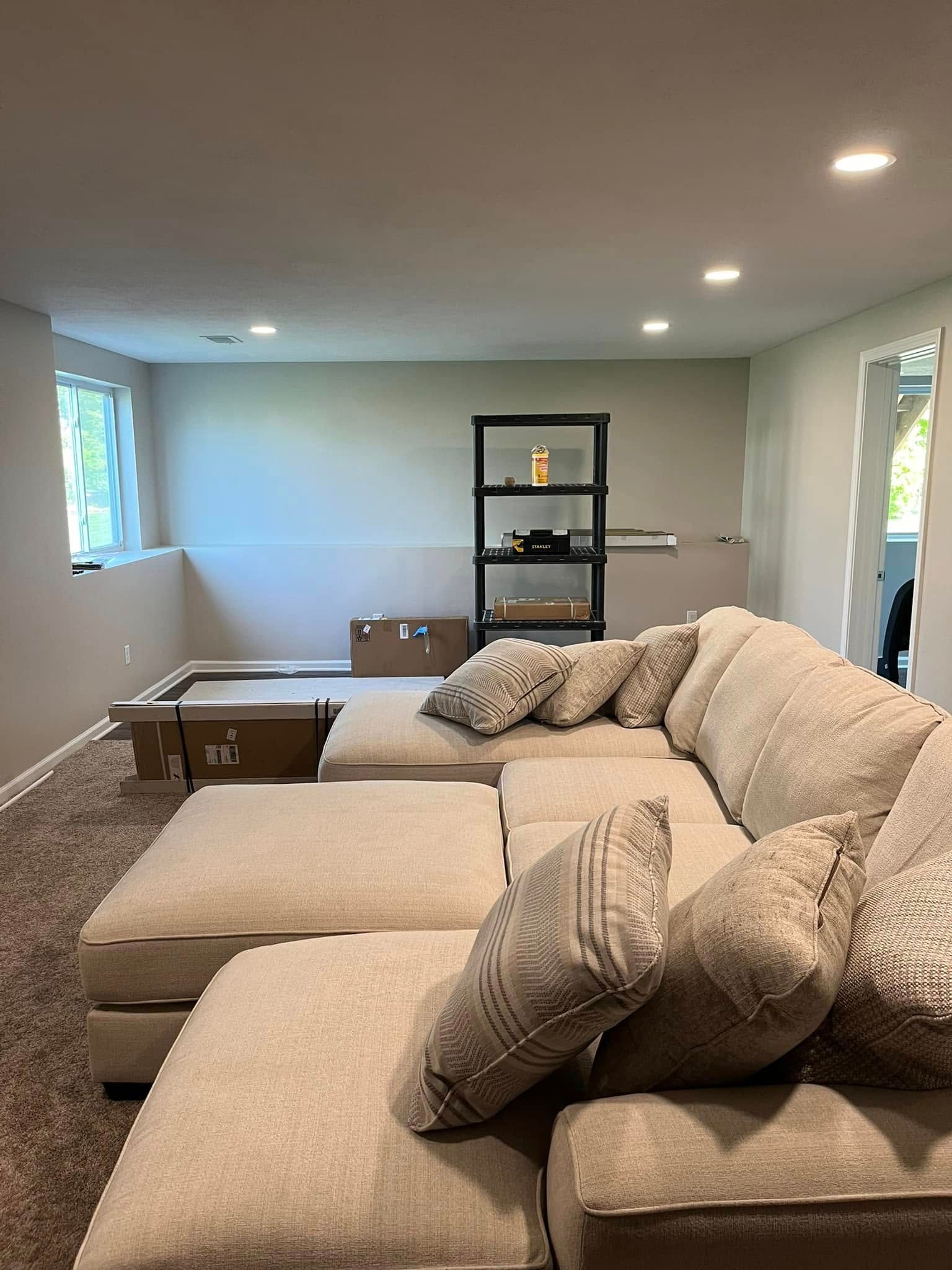 Basement living room with beige sectional sofa, ottoman, coffee table, and shelving unit