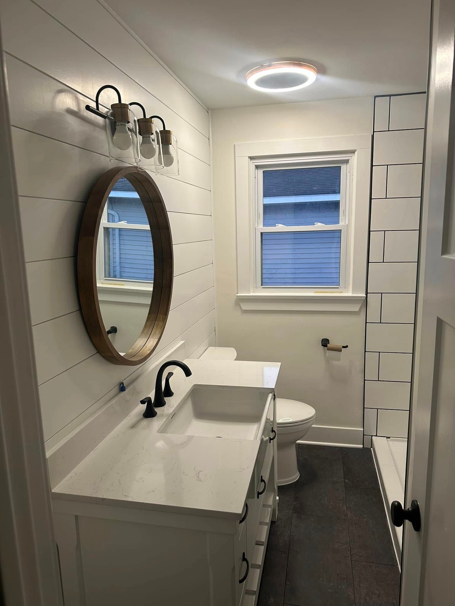 Narrow white bathroom with oval mirror, black faucet, window, and shower curtain on the right.