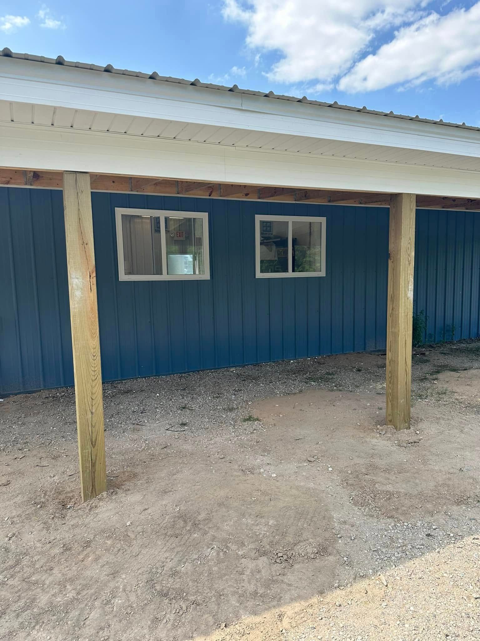 Blue building with two windows, white roof, and two wooden support posts on gravel under a blue sky