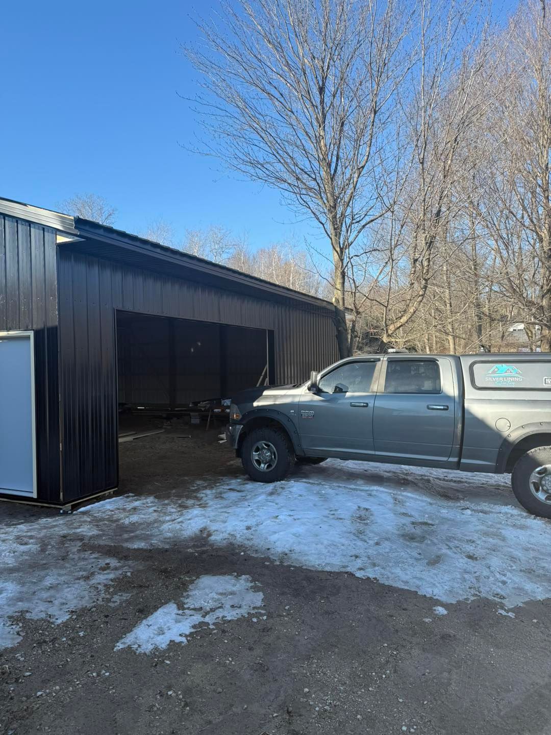 Gray pickup truck parked beside a dark metal garage on a snowy driveway under a clear blue sky