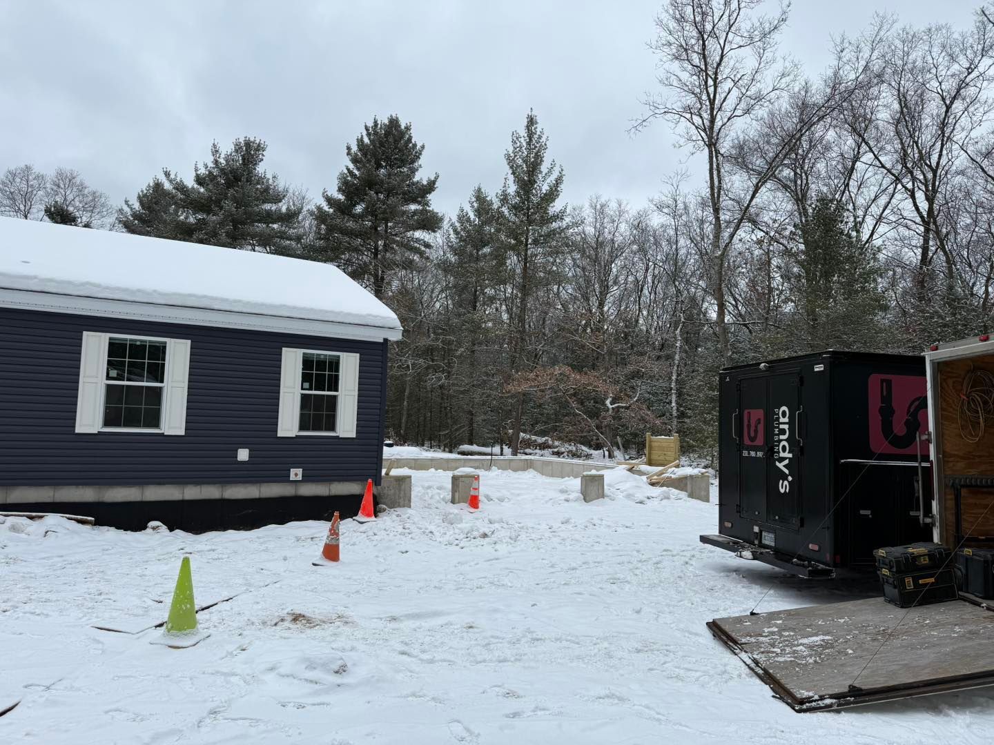 Snowy yard beside a dark house, with orange cones and a black trailer near leafless trees.