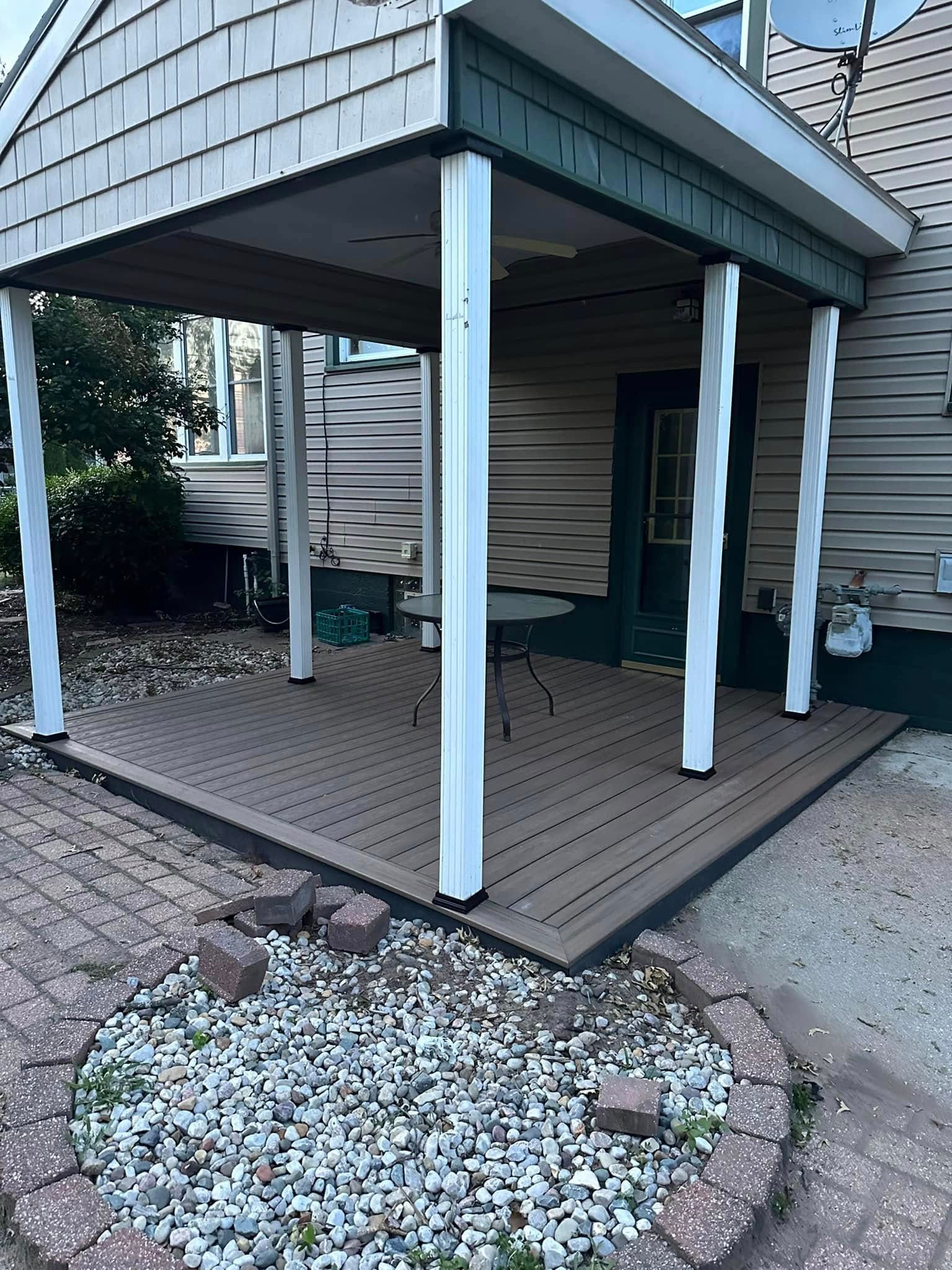 Covered wooden deck with white posts and railing beside a house, bordered by gravel and brick edging
