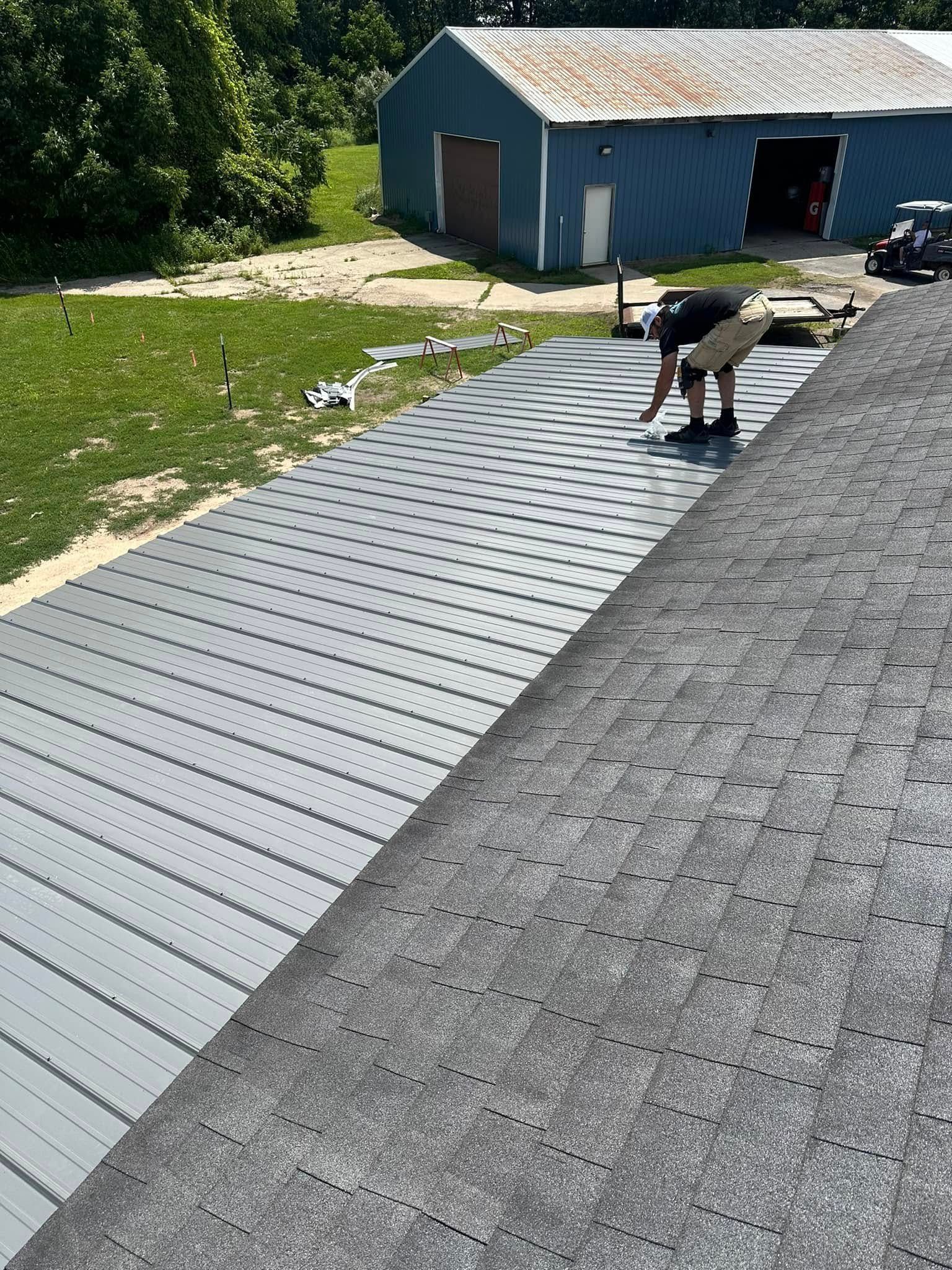Worker installing silver metal roofing beside a shingled roof on a house under construction
